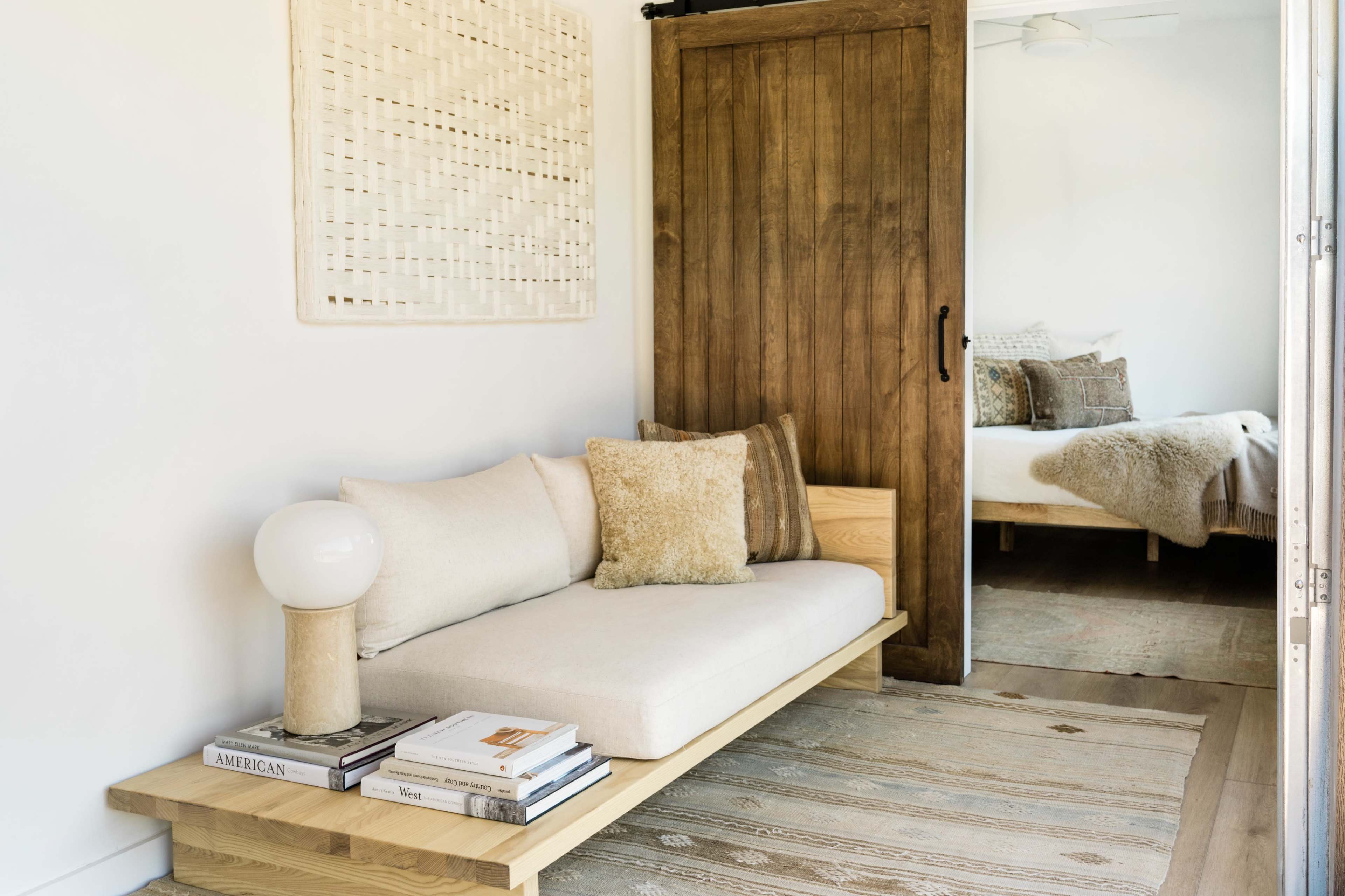 A minimalist seating area features a light-colored sofa with throw pillows and a lamp, next to a wooden barn door leading to a bedroom.