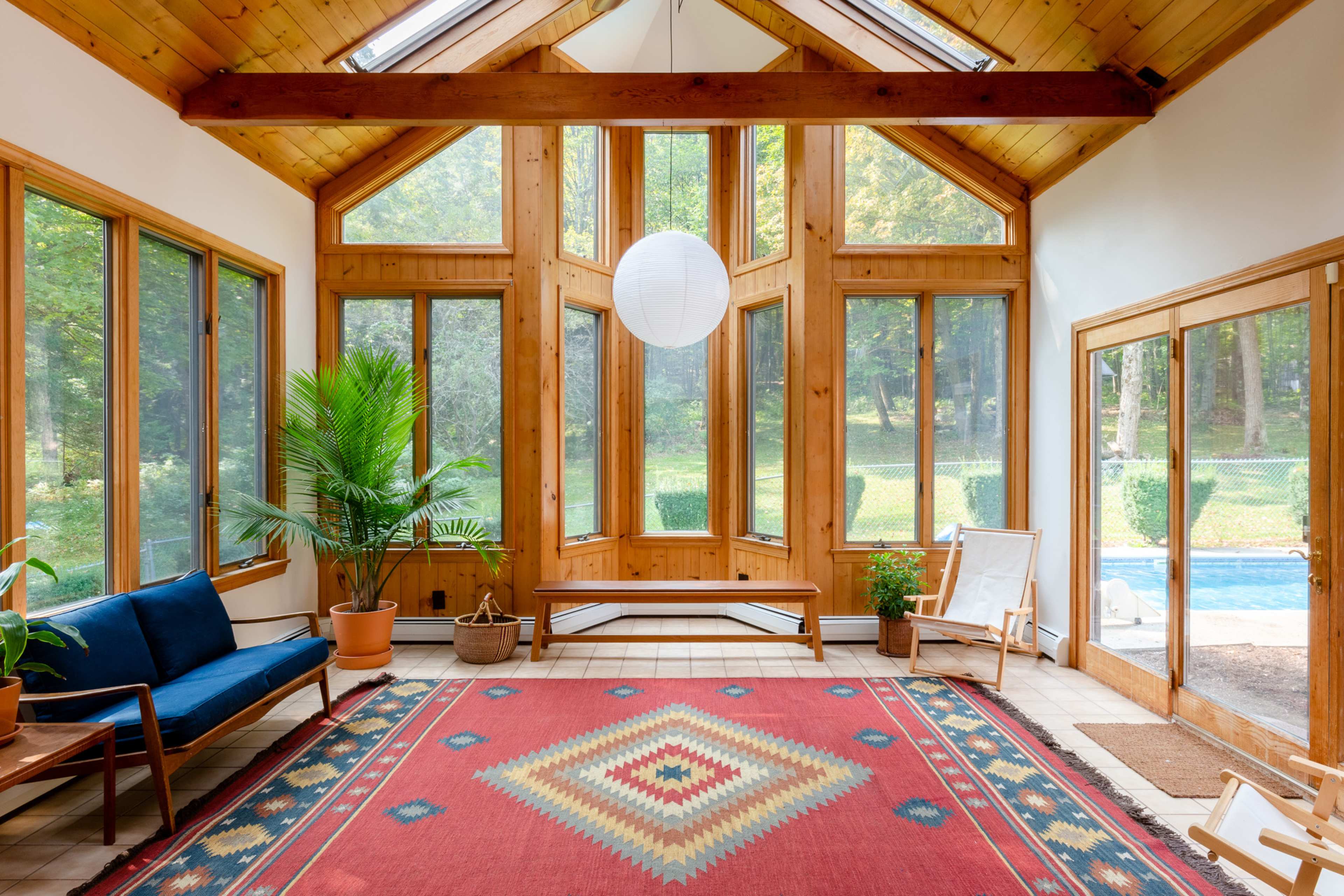 A sunlit sunroom features large windows, wooden beams, a red area rug with a geometric pattern, and plants surrounding a seating area.