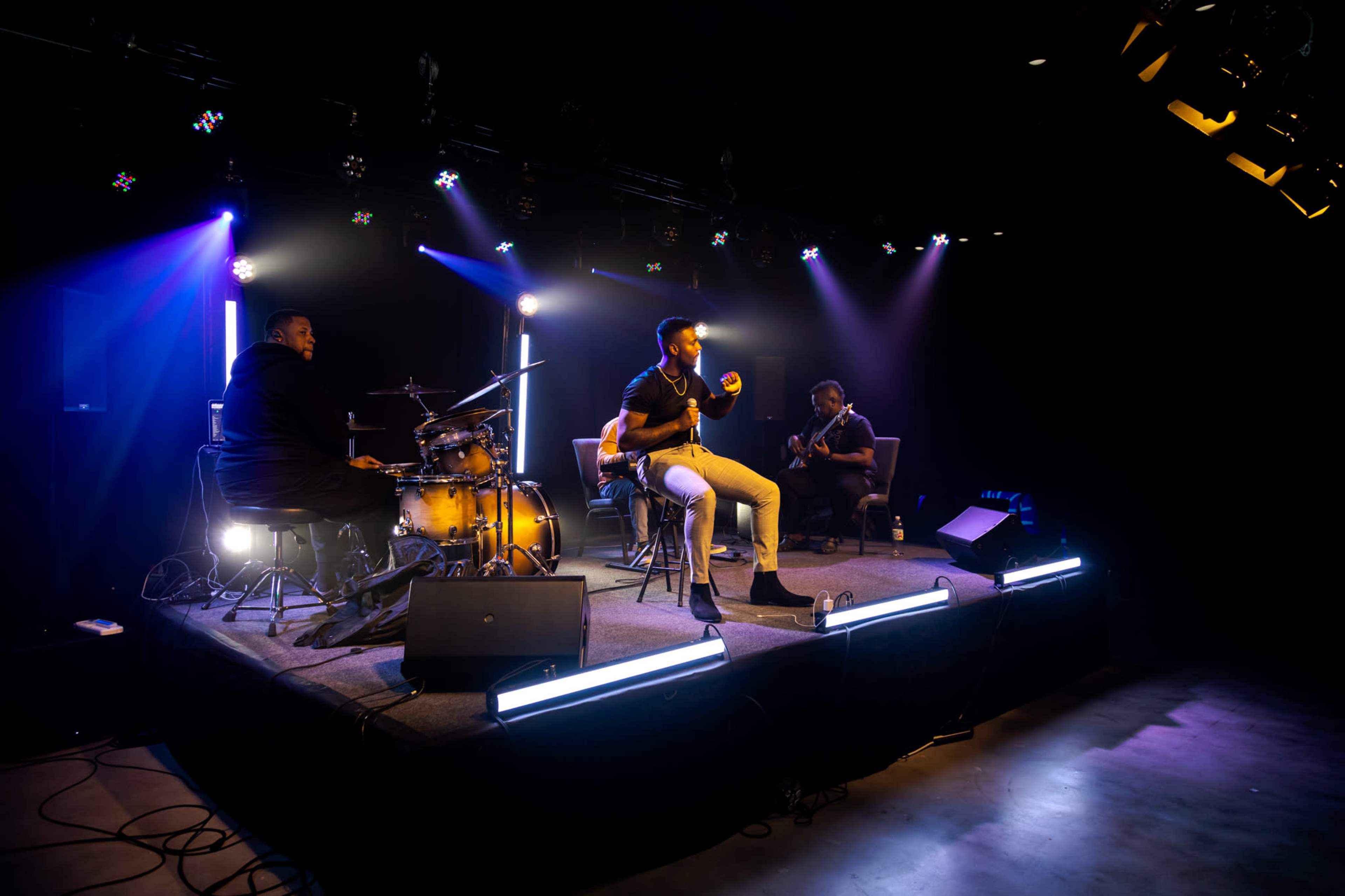 A musician performs on stage with a drummer and another guitarist in a dimly lit venue illuminated by stage lights.