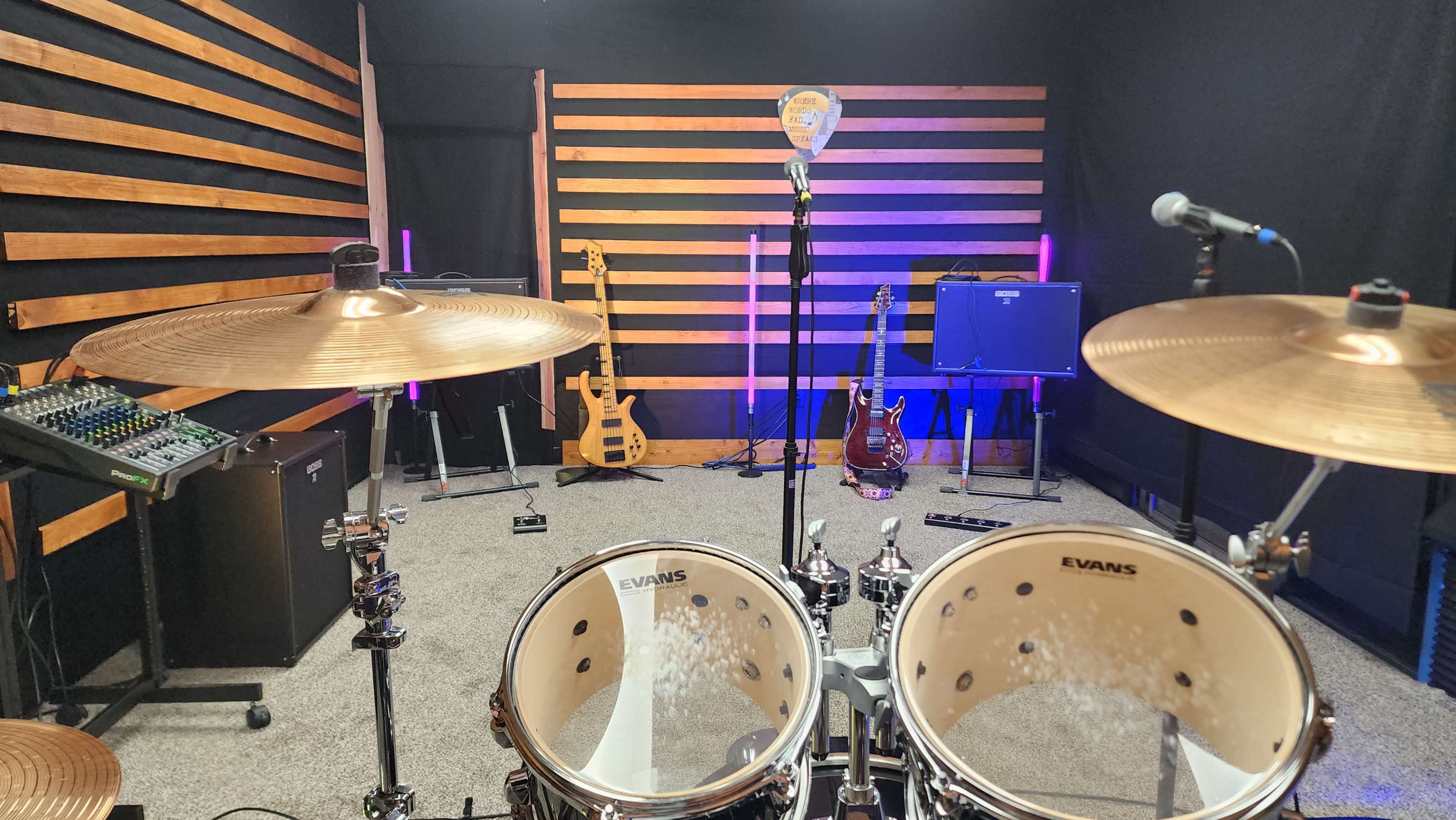 The image shows a music practice room with a drum set in the foreground, a microphone stand, and various guitars displayed on the wall.