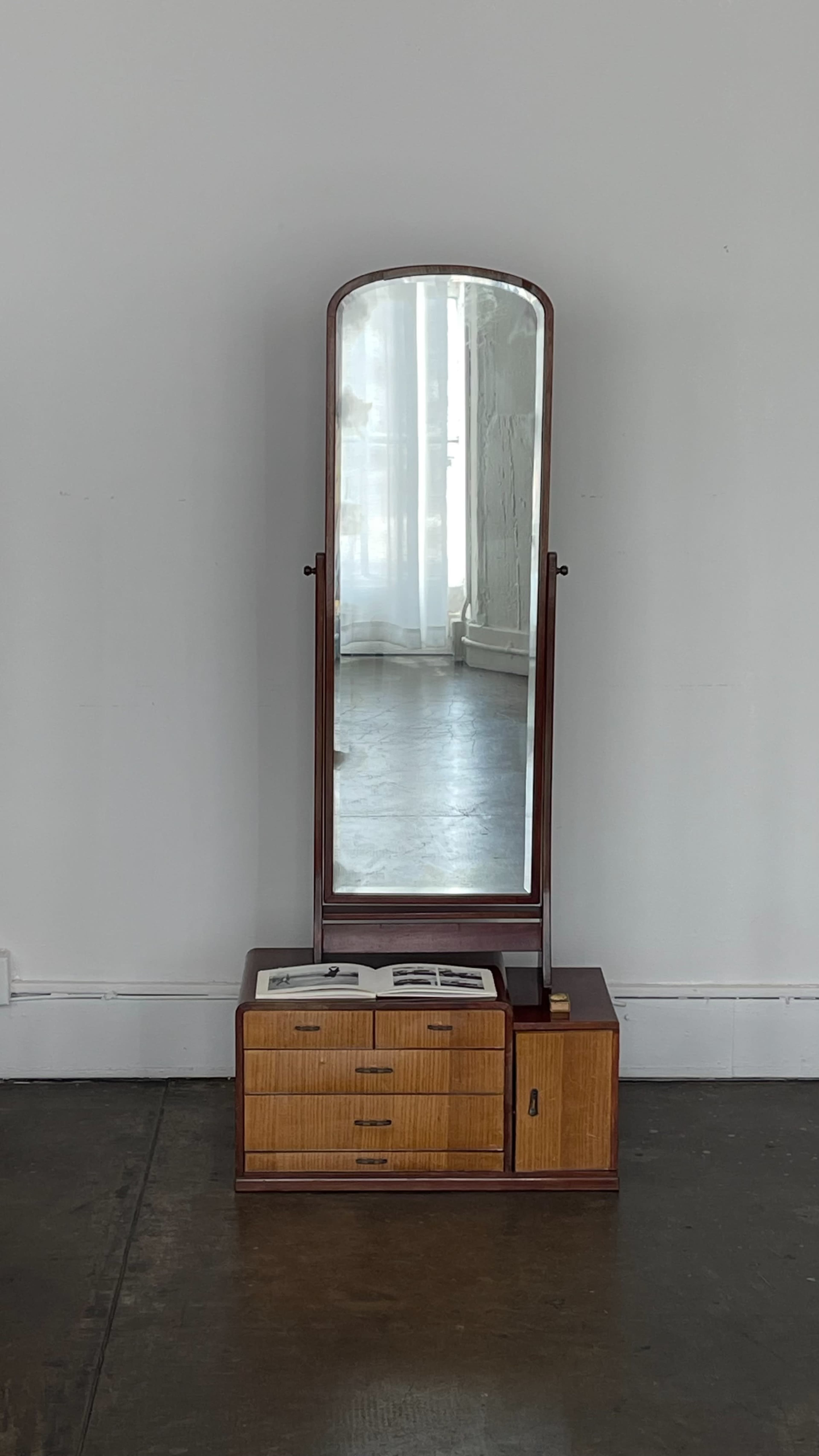 A tall mirror stands on a wooden dresser with drawers, positioned against a plain wall in a well-lit room.