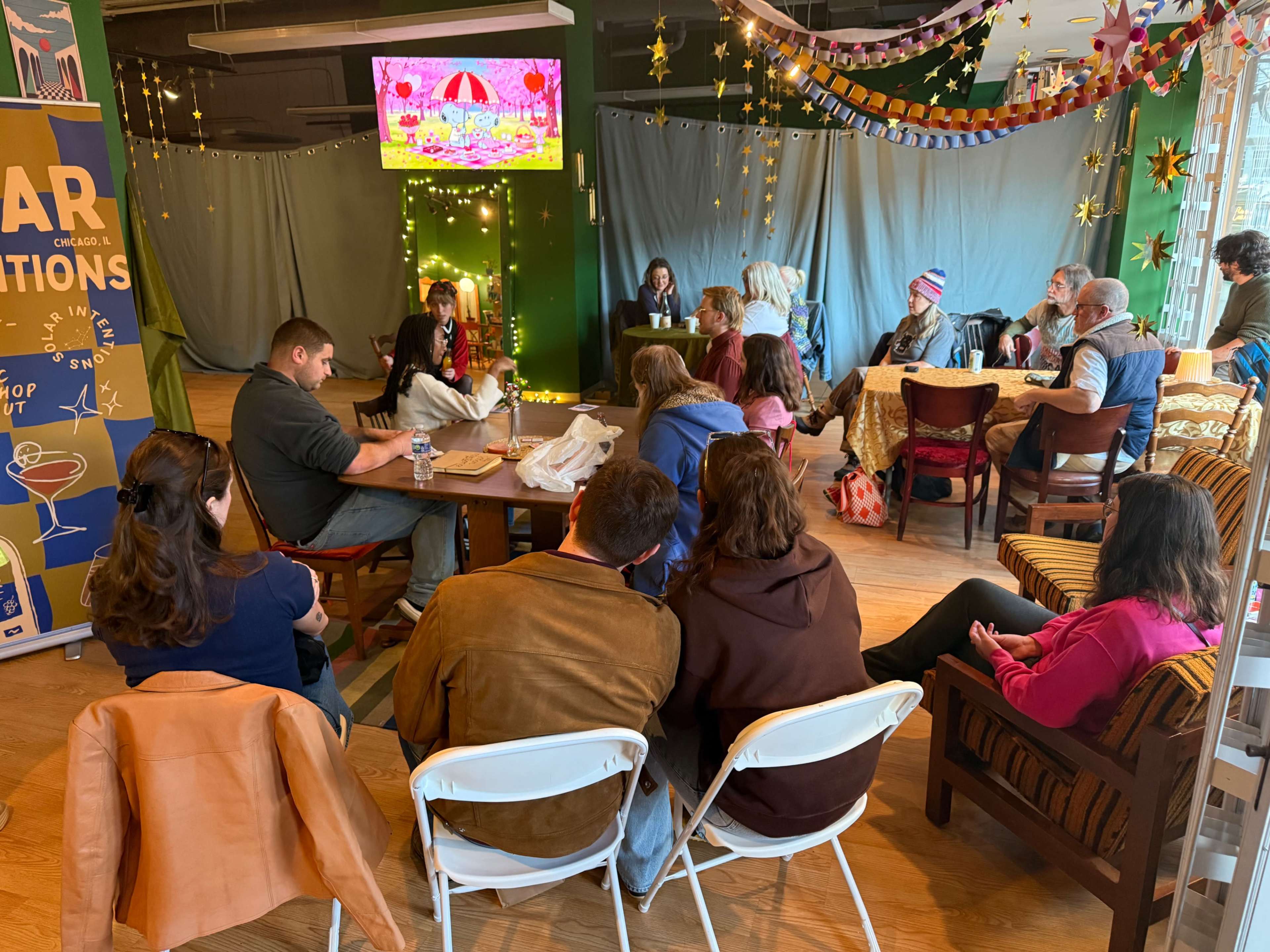 A group of people sits in a cozy venue with decorations and a screen displaying an animated image, while some engage in conversation and others focus on the presentation.