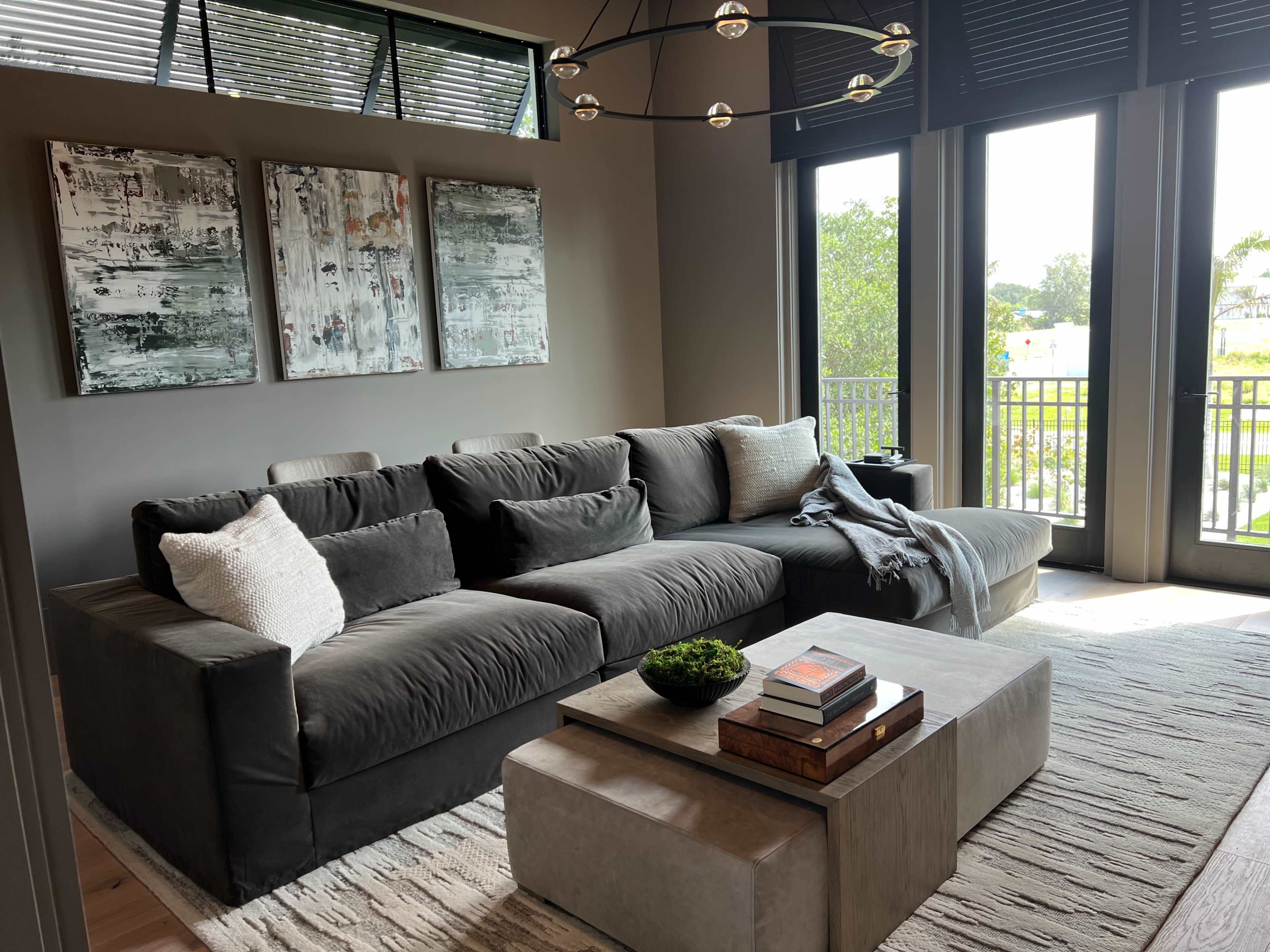 A spacious living room features a large sectional sofa, a coffee table with stacked books and a small plant, and large windows with natural light.