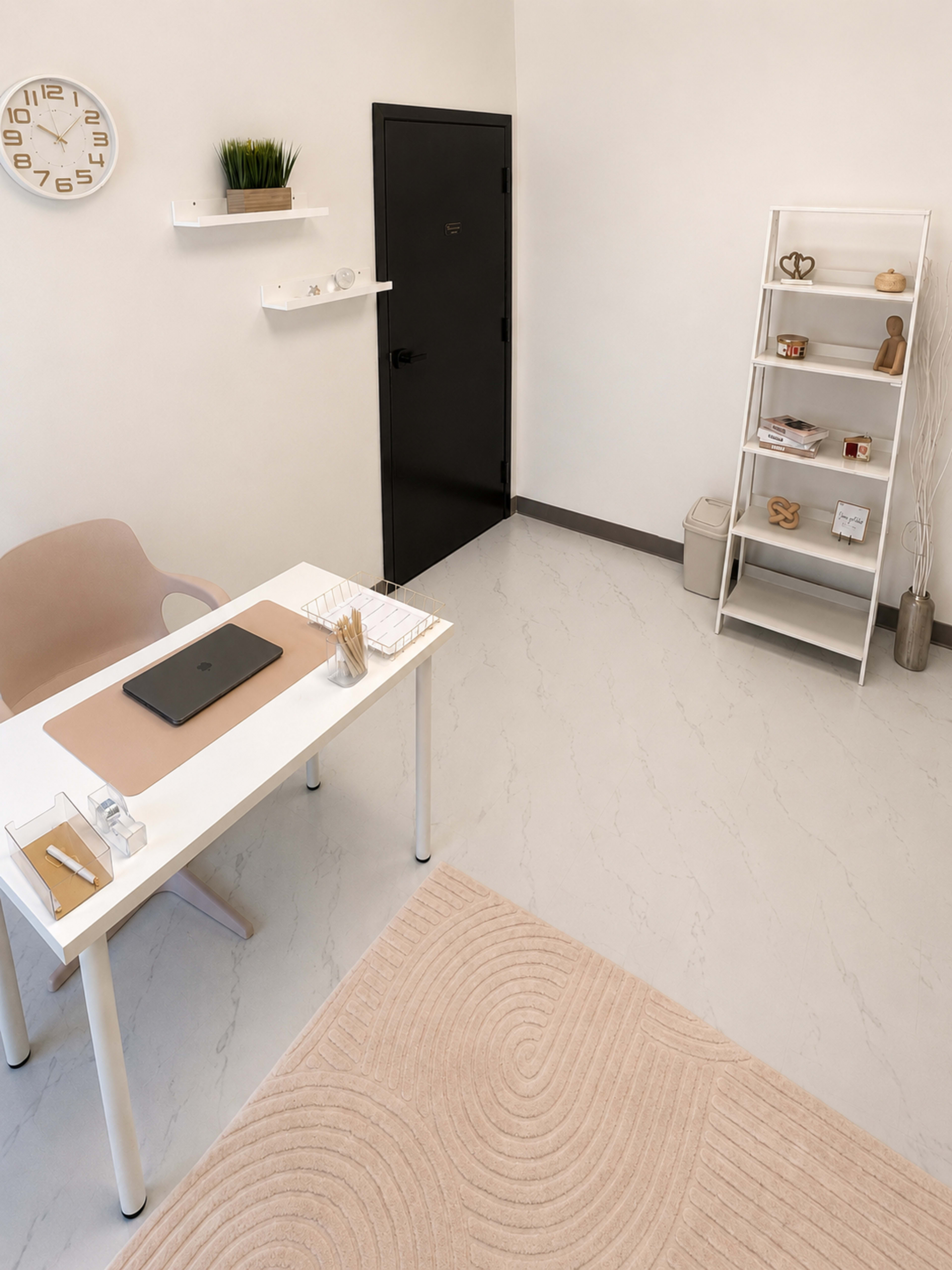The image shows a modern office space featuring a desk with a laptop, a chair, a shelf with decorative items, and a door leading to another room.
