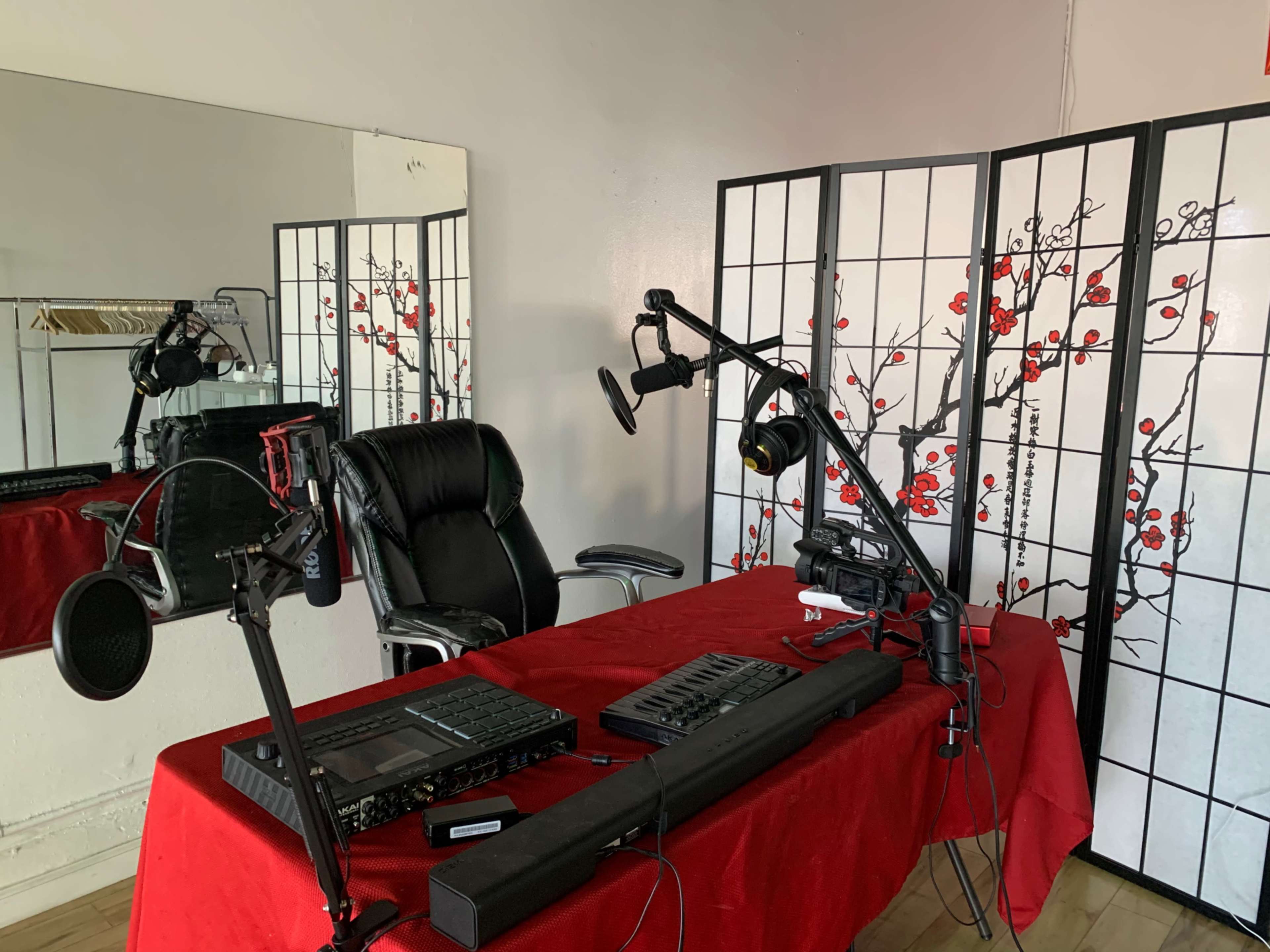 A recording studio setup with a microphone, audio equipment, and a chair positioned in front of a red tablecloth and a decorative screen.