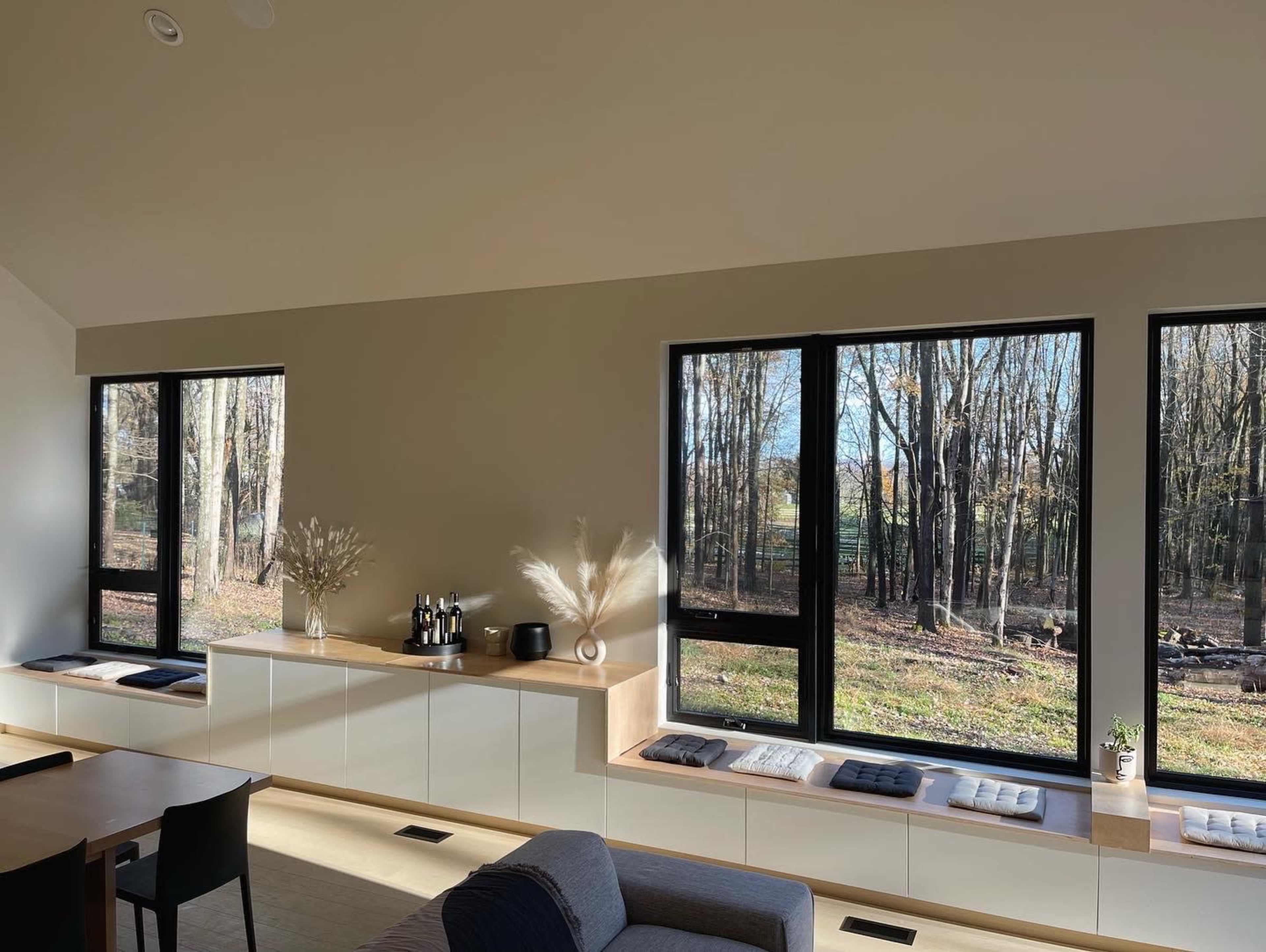A modern interior features large windows overlooking a wooded area, with minimalist furniture and decor.