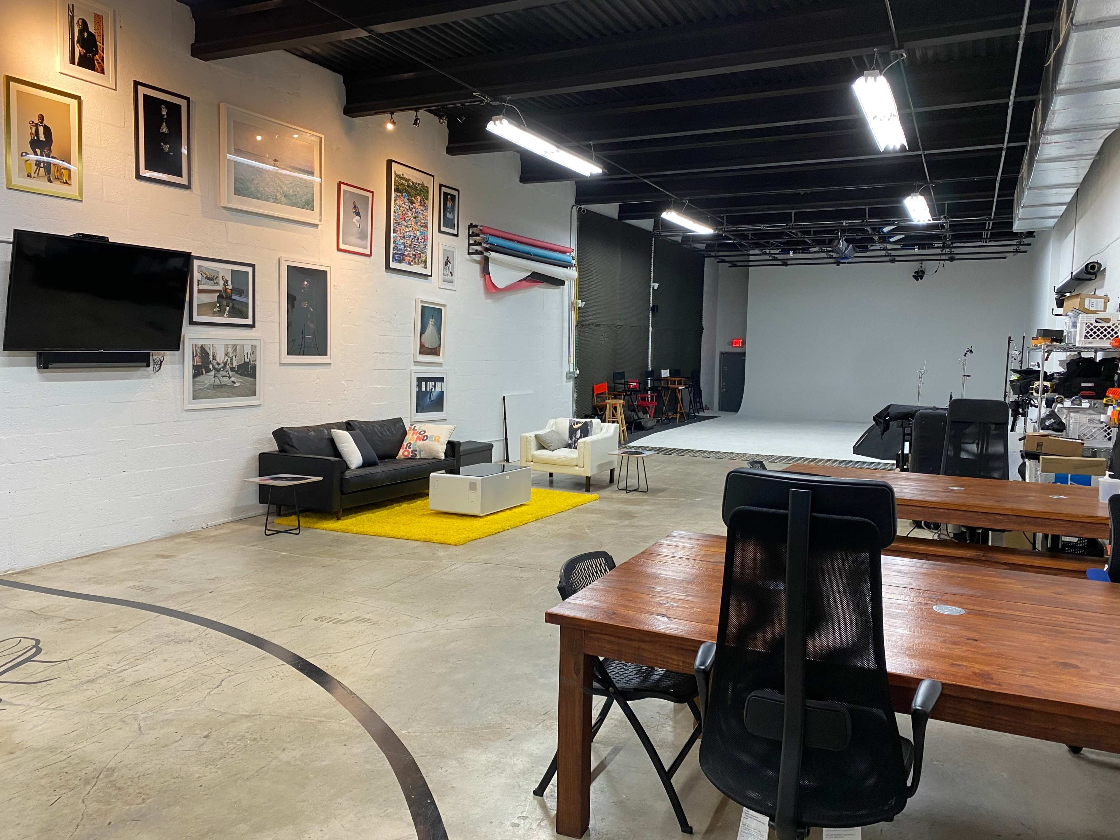 A spacious studio featuring a black sofa, a white armchair, a yellow rug, and a large backdrop area with various studio equipment and framed photographs on the walls.