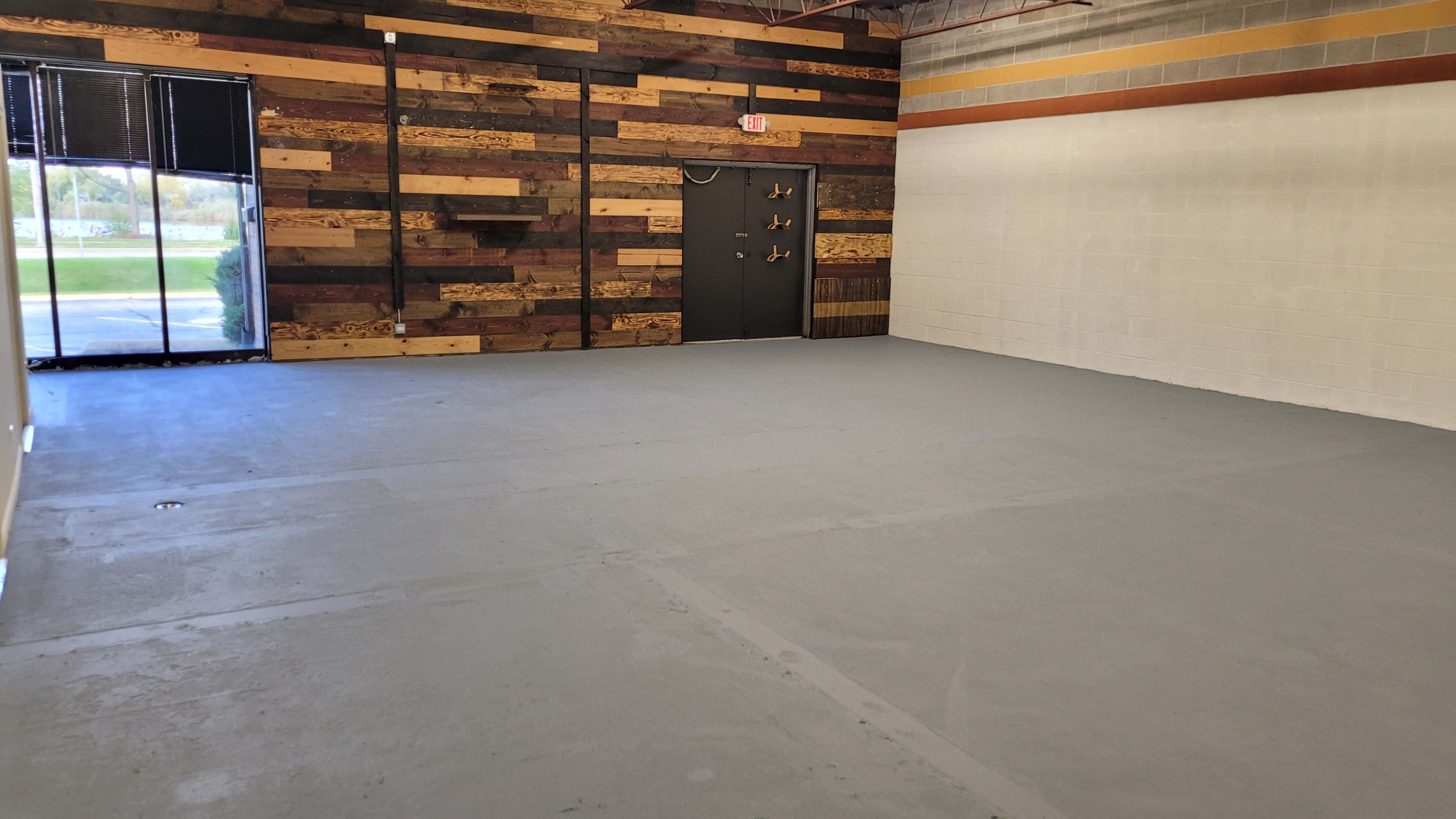 An empty room with a concrete floor, a dark door on one wall, and a wood-clad wall featuring various tones of wood.