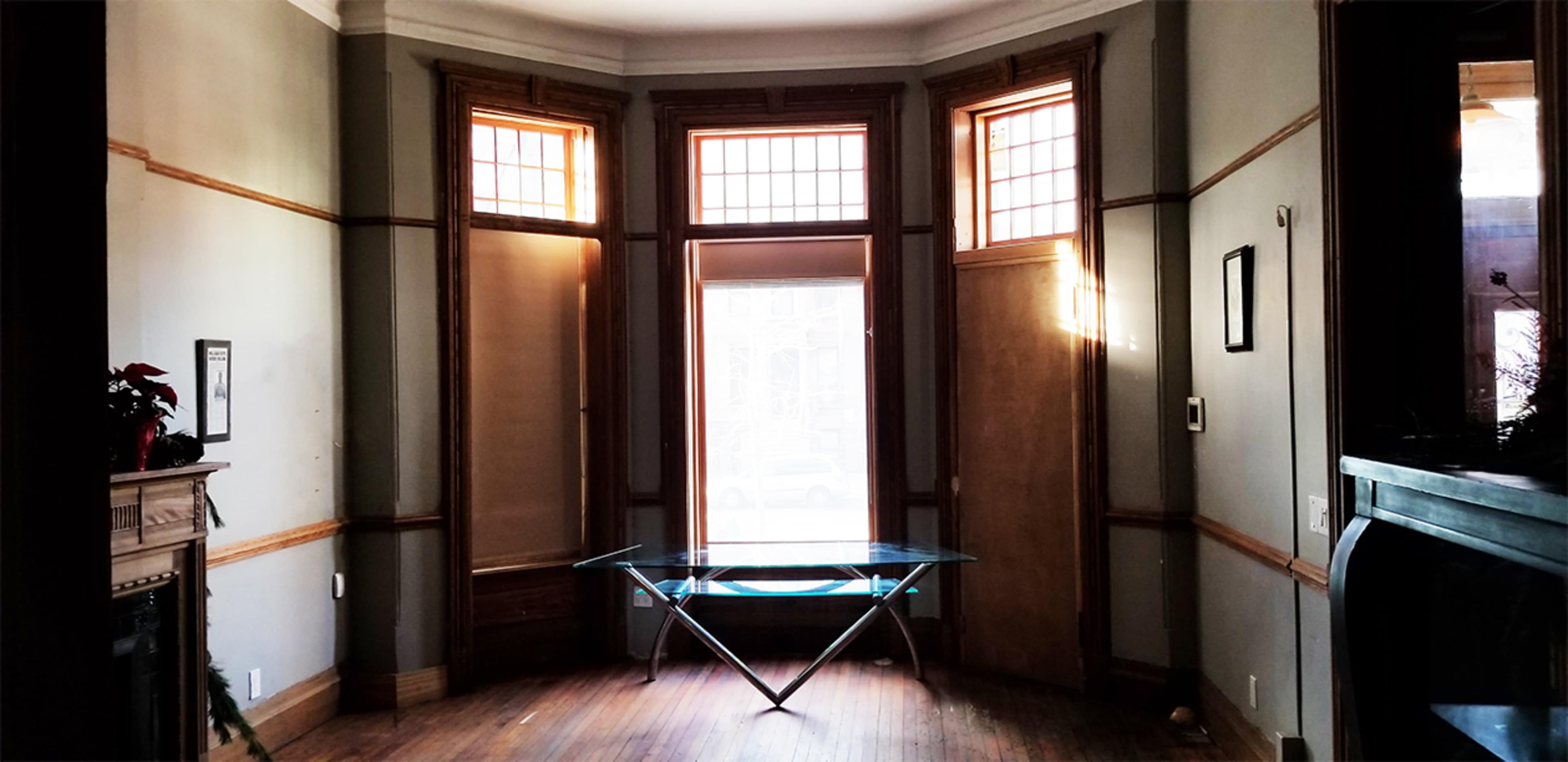 The image shows a room with large windows flanked by wooden trim and a glass table in the center.