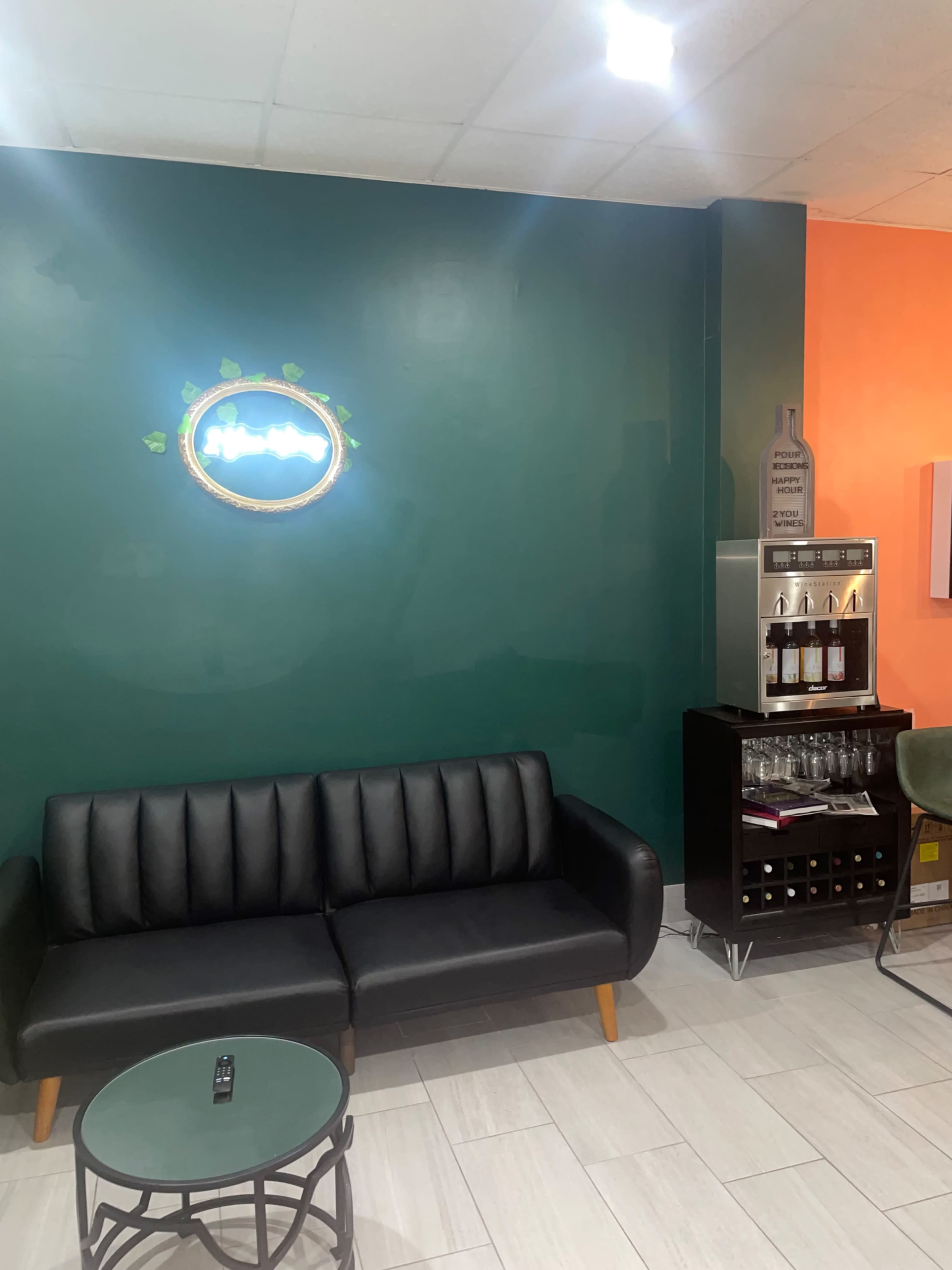 The image shows a small lounge area with a black leather sofa, a circular coffee table, and a beverage station against a green wall.