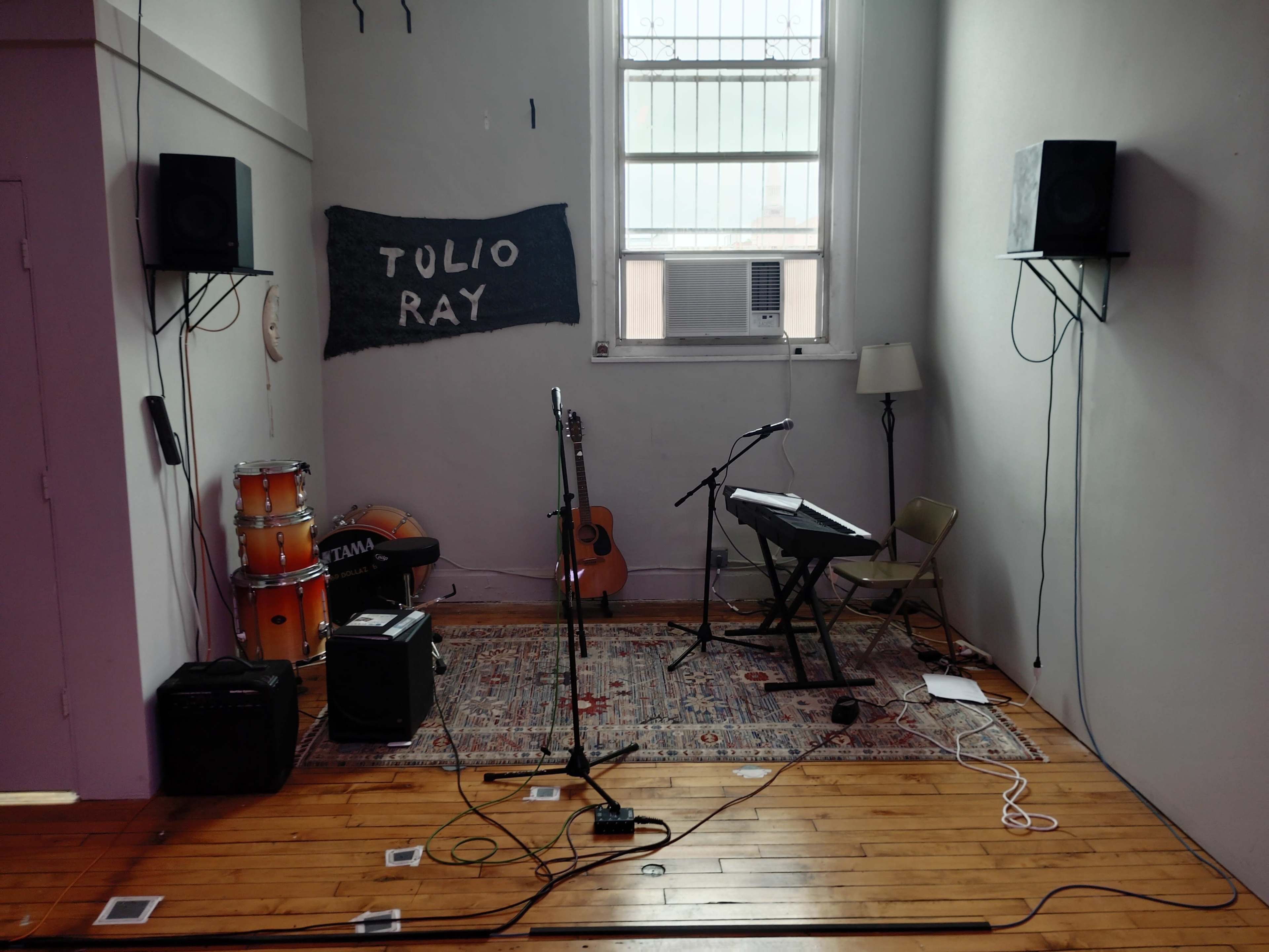 The image shows an empty room set up for a music performance, featuring instruments like a drum set, guitar, keyboard, and amplifiers along with a carpet and a window.