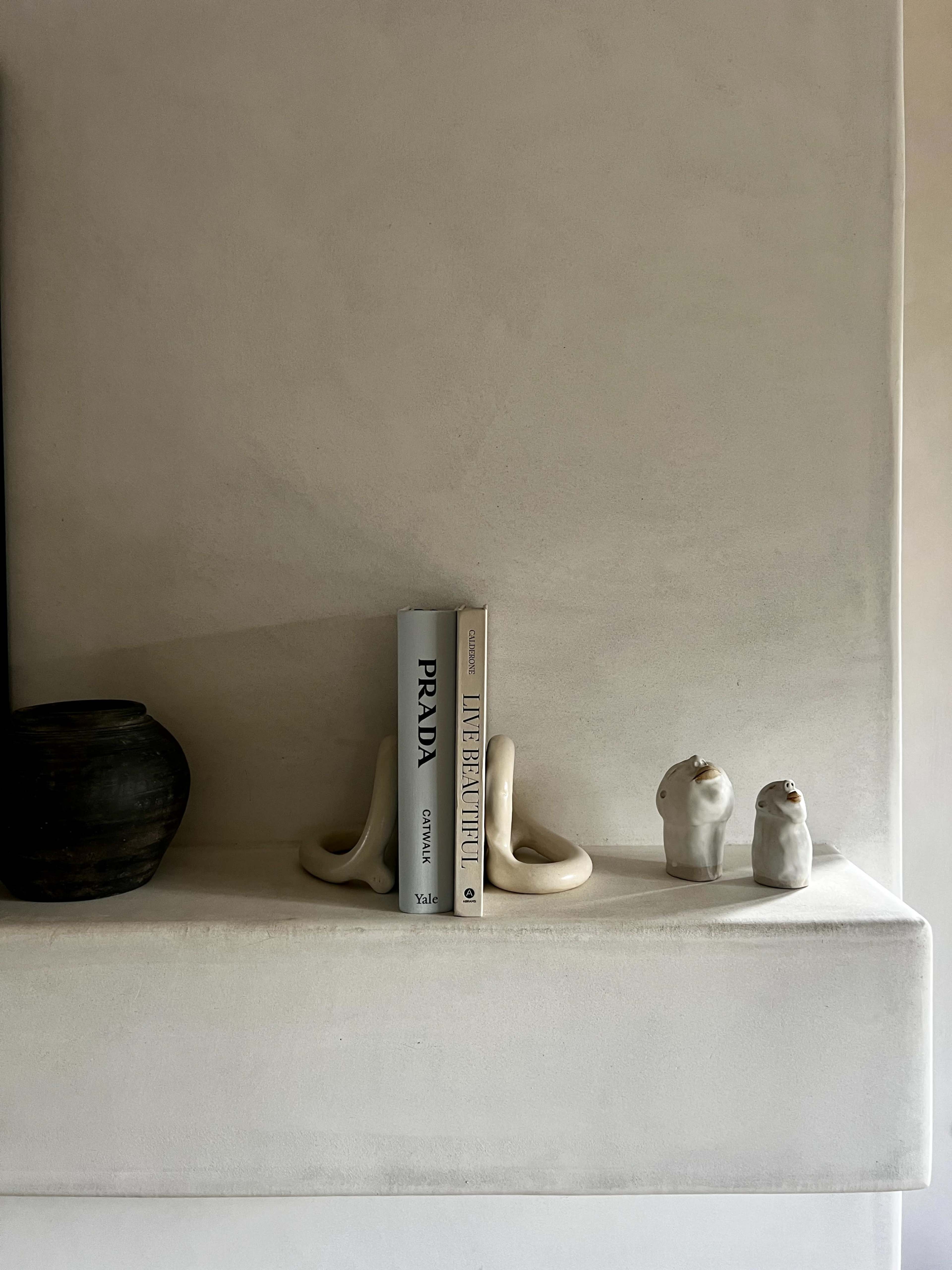 A minimalist shelf displays a black ceramic vase, a book titled "Prada," and two small white sculptures against a textured wall.