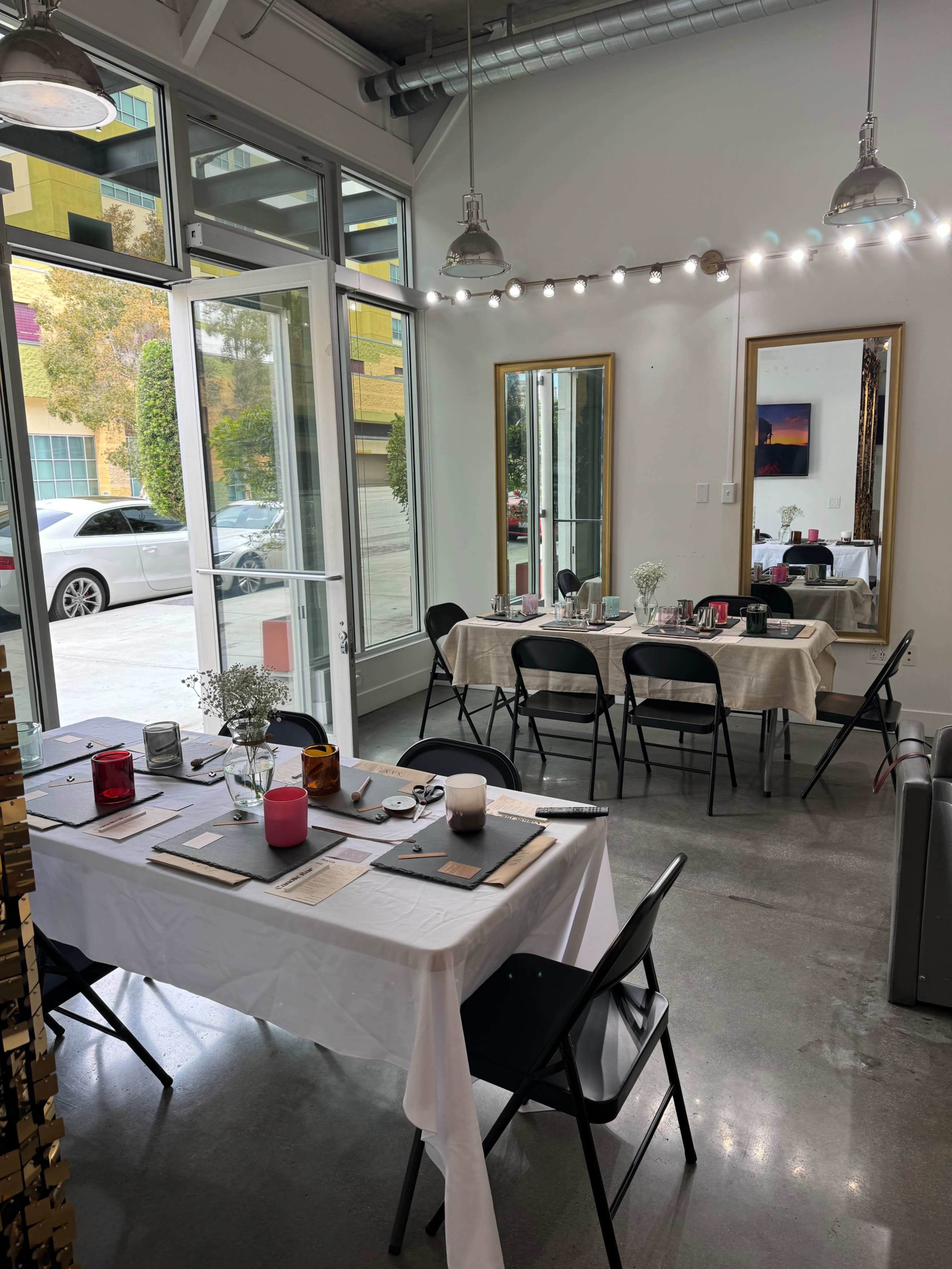 A modern, bright room set up for dining, featuring white tables with tablecloths, black chairs, and decorative mirrors, with large windows allowing natural light to enter.
