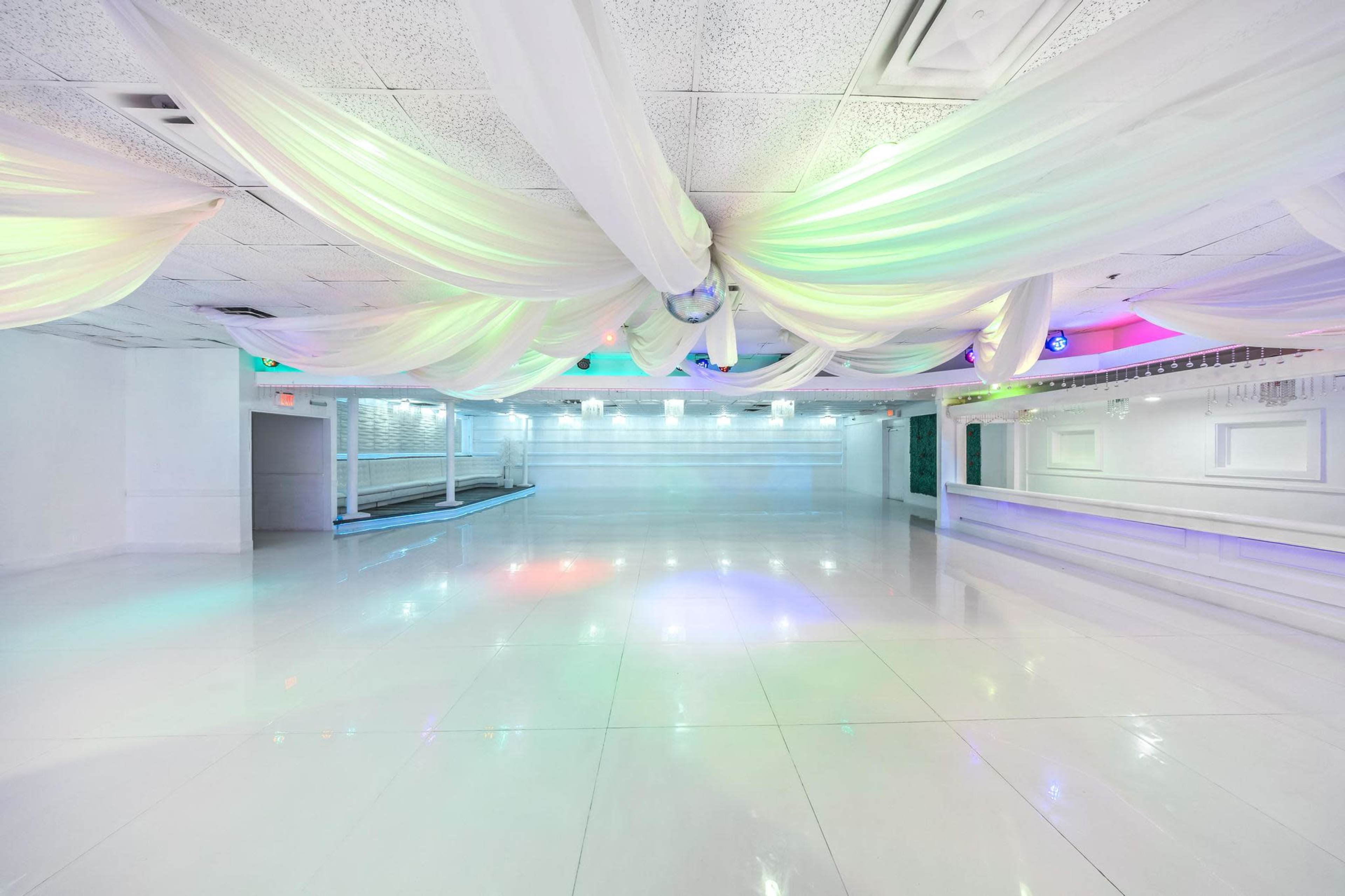 The image shows a spacious indoor event venue with white walls and a glossy floor, featuring decorative fabric draping from the ceiling and colored LED lights illuminating the room.