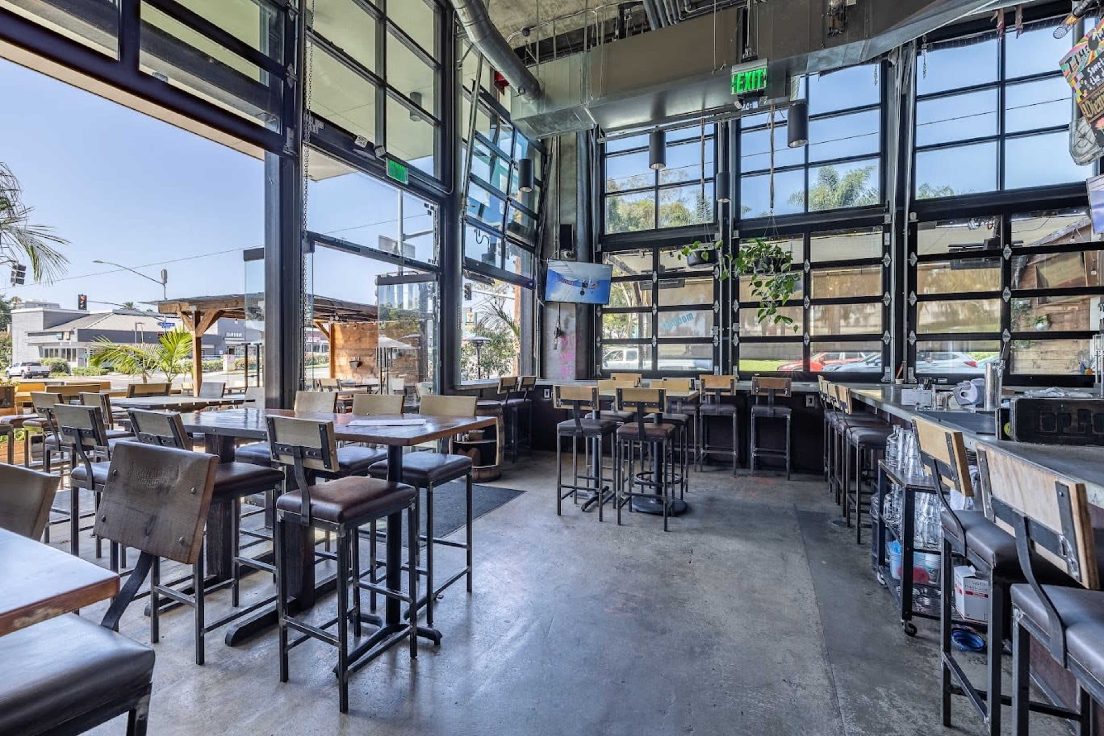 The interior of a modern restaurant with large windows, high tables, and bar stools arranged around an open space.