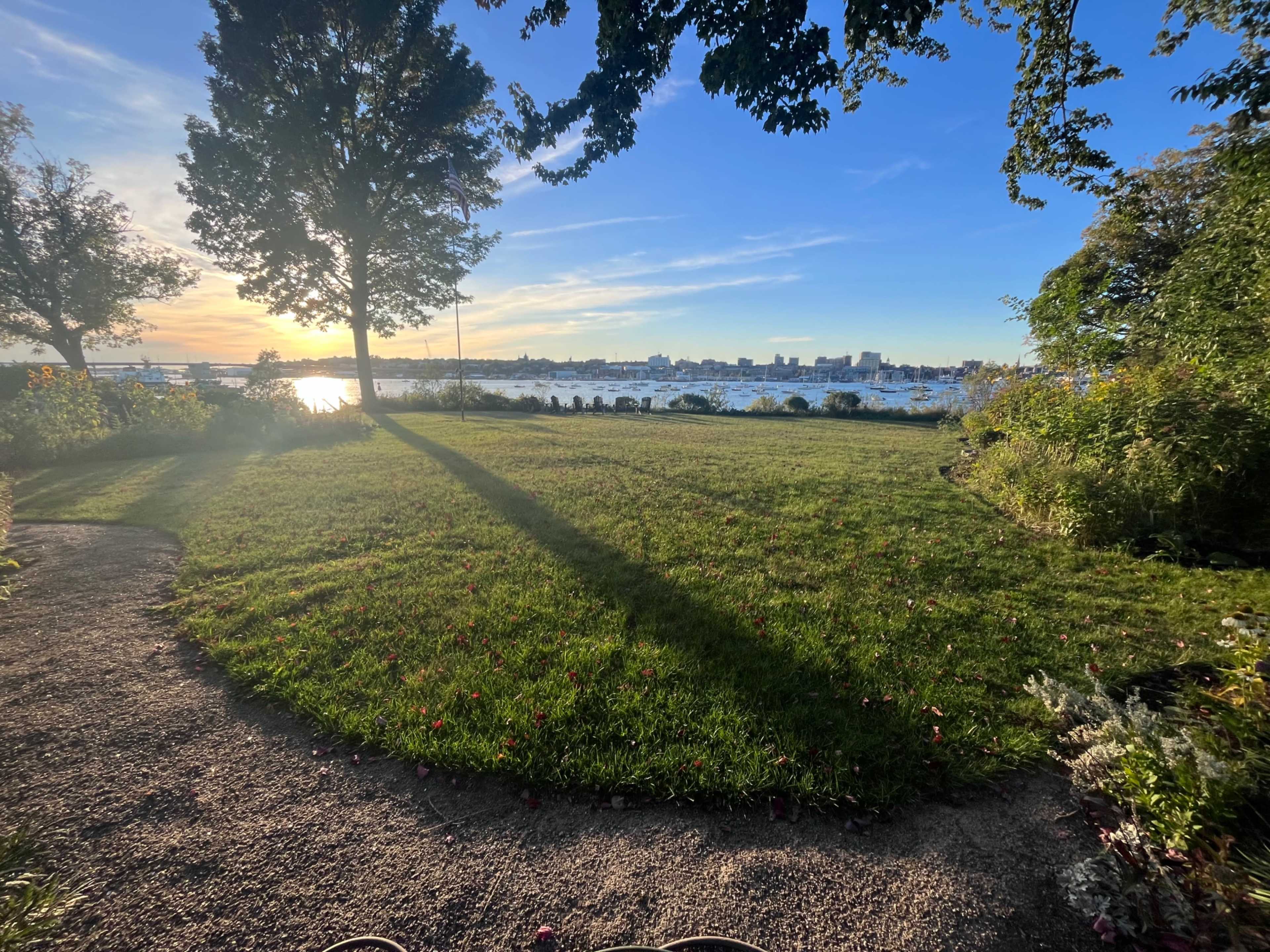 The image shows a grassy area with a path leading towards a waterfront view at sunset, framed by trees and flower gardens.