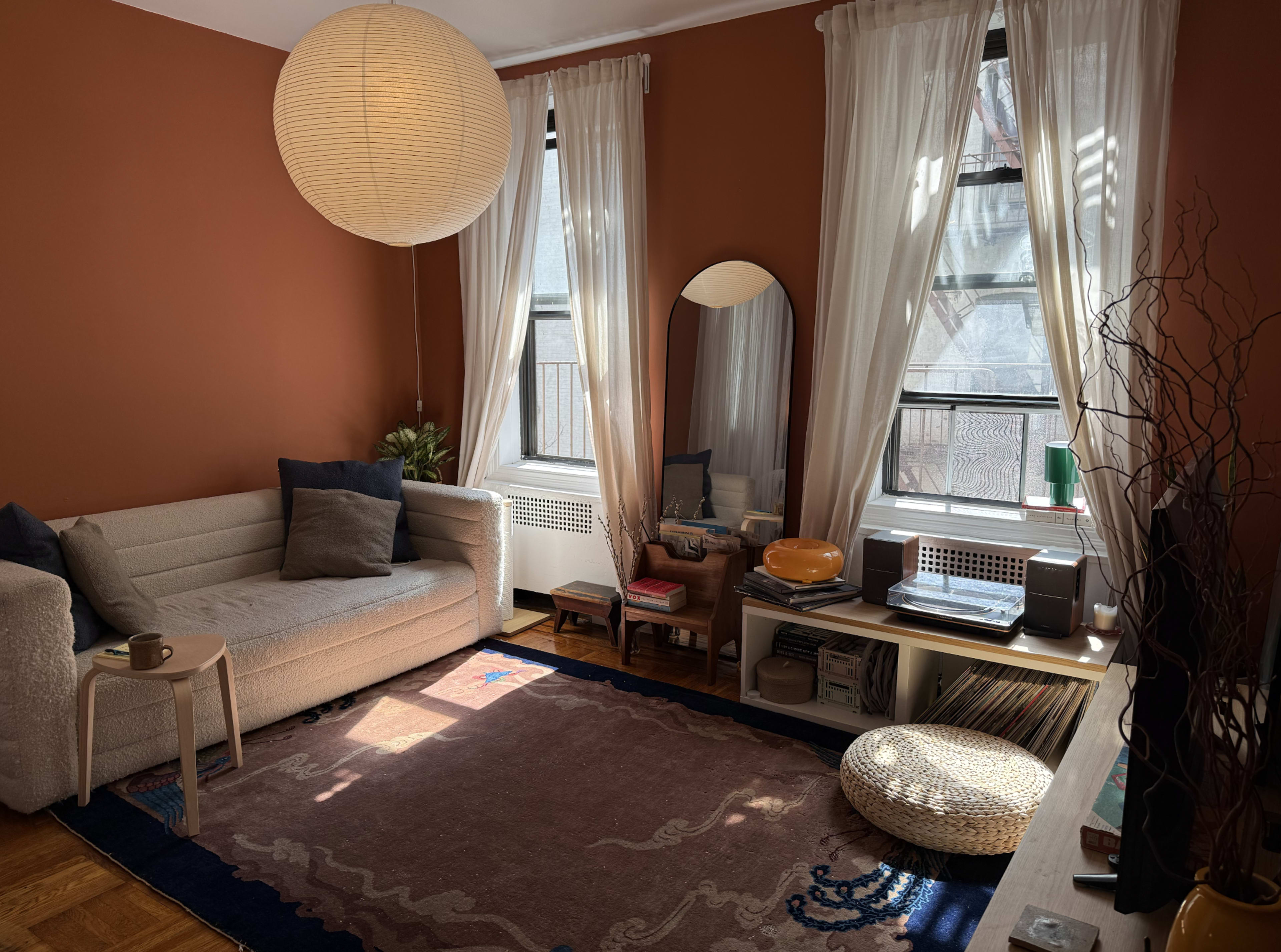 The image shows a cozy living room with a beige sofa, a round paper lantern, large windows covered with sheer curtains, and a patterned rug on wooden flooring.