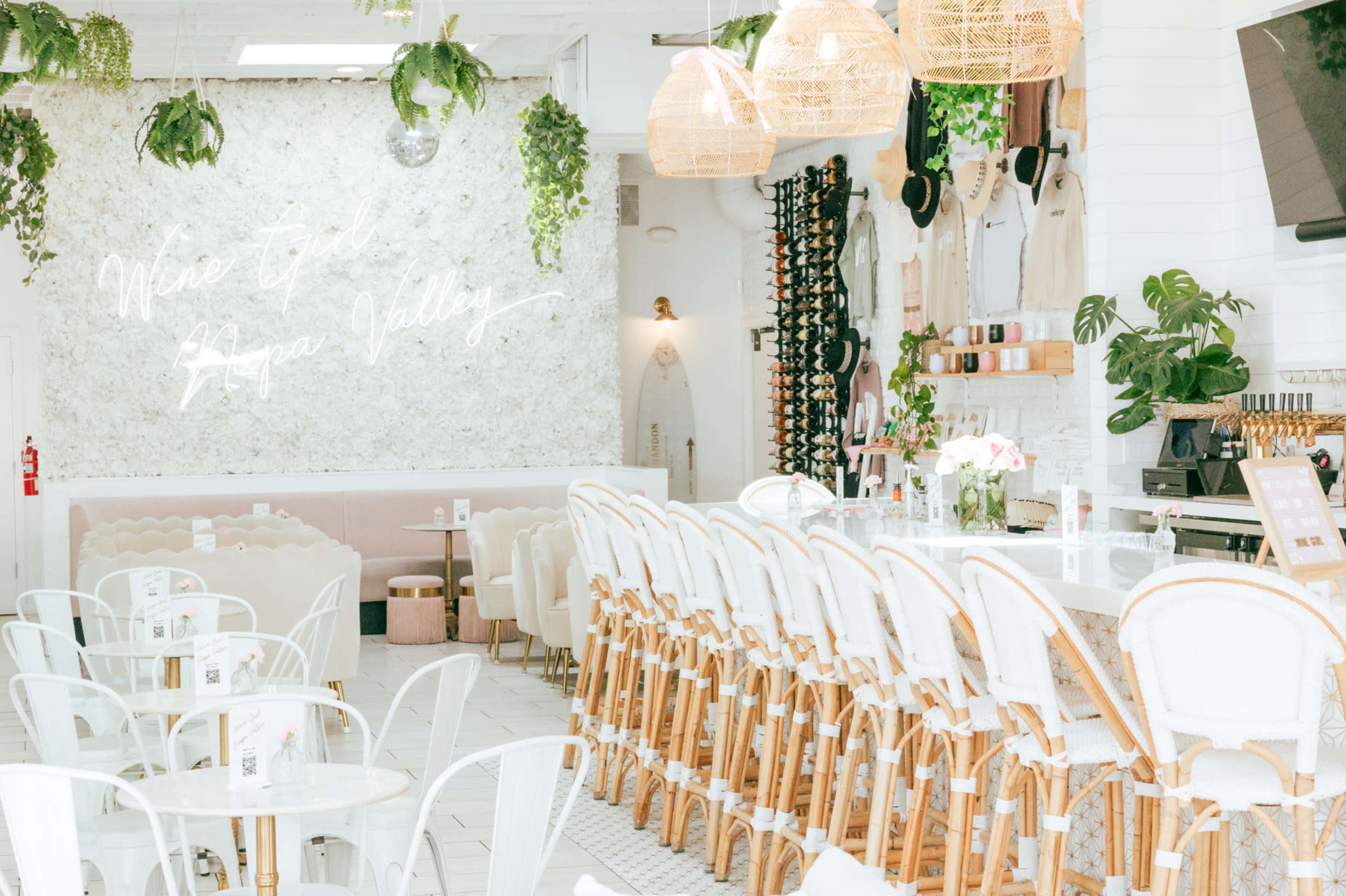The image shows a bright, stylish café featuring white furniture, a bar with high stools, and greenery hanging from the ceiling, along with a neon sign on the wall.