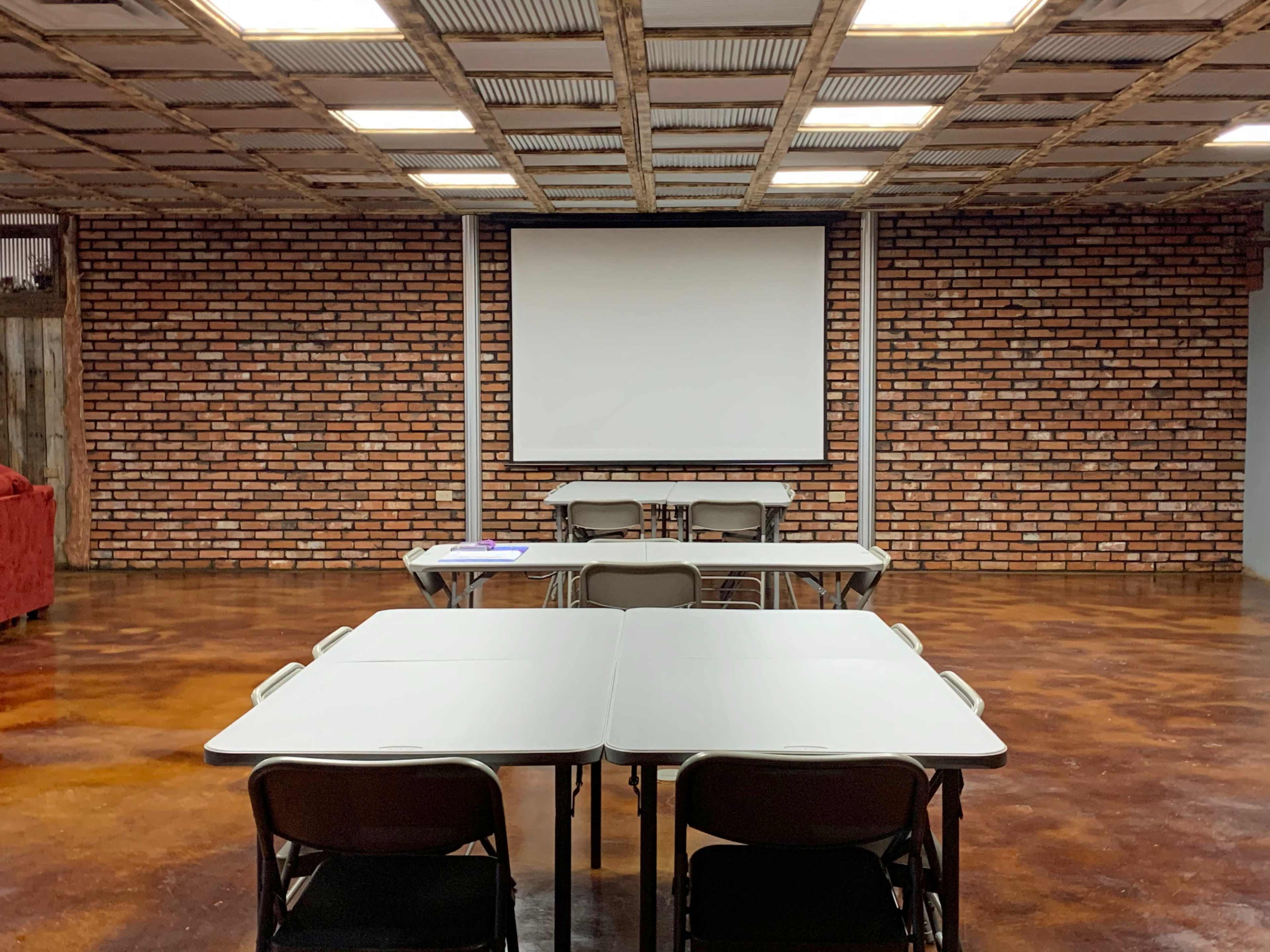 A room with a brick wall, a large projection screen, and several tables and chairs arranged in a meeting setup.