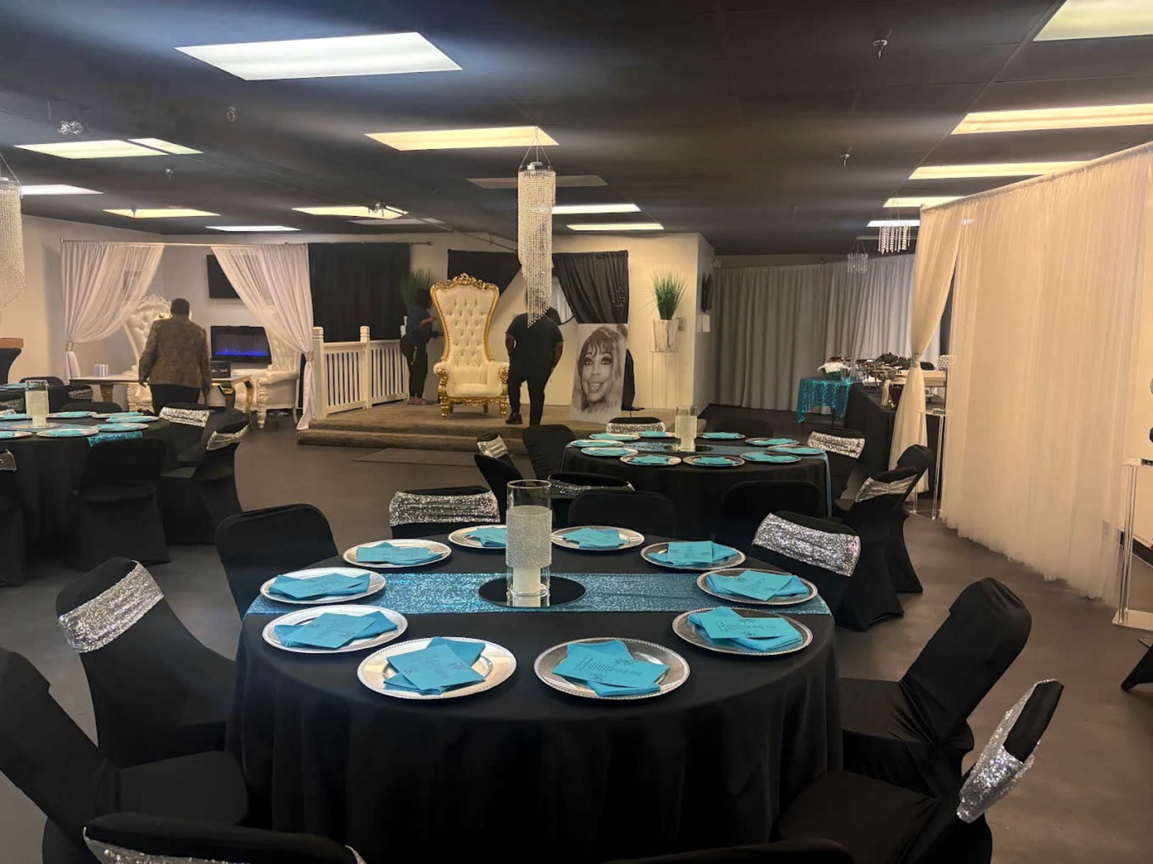 The image shows a banquet hall setup with several round tables covered in black cloth, each adorned with turquoise plates and silver accents, while a large, ornate chair is displayed against a backdrop featuring a large portrait.