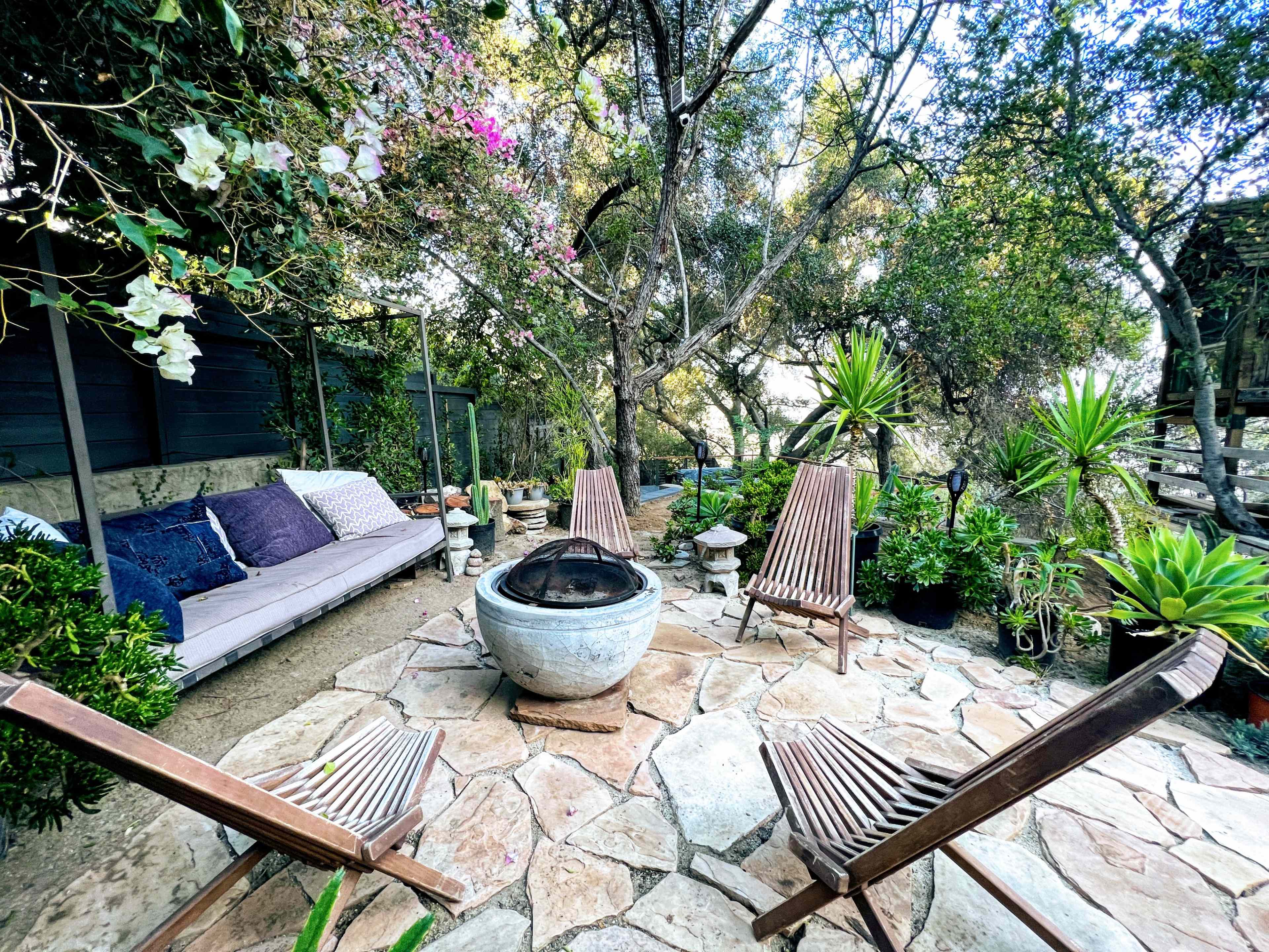 A stone patio features a circular fire pit surrounded by wooden lounge chairs and lush greenery, with a sofa draped with cushions nearby.