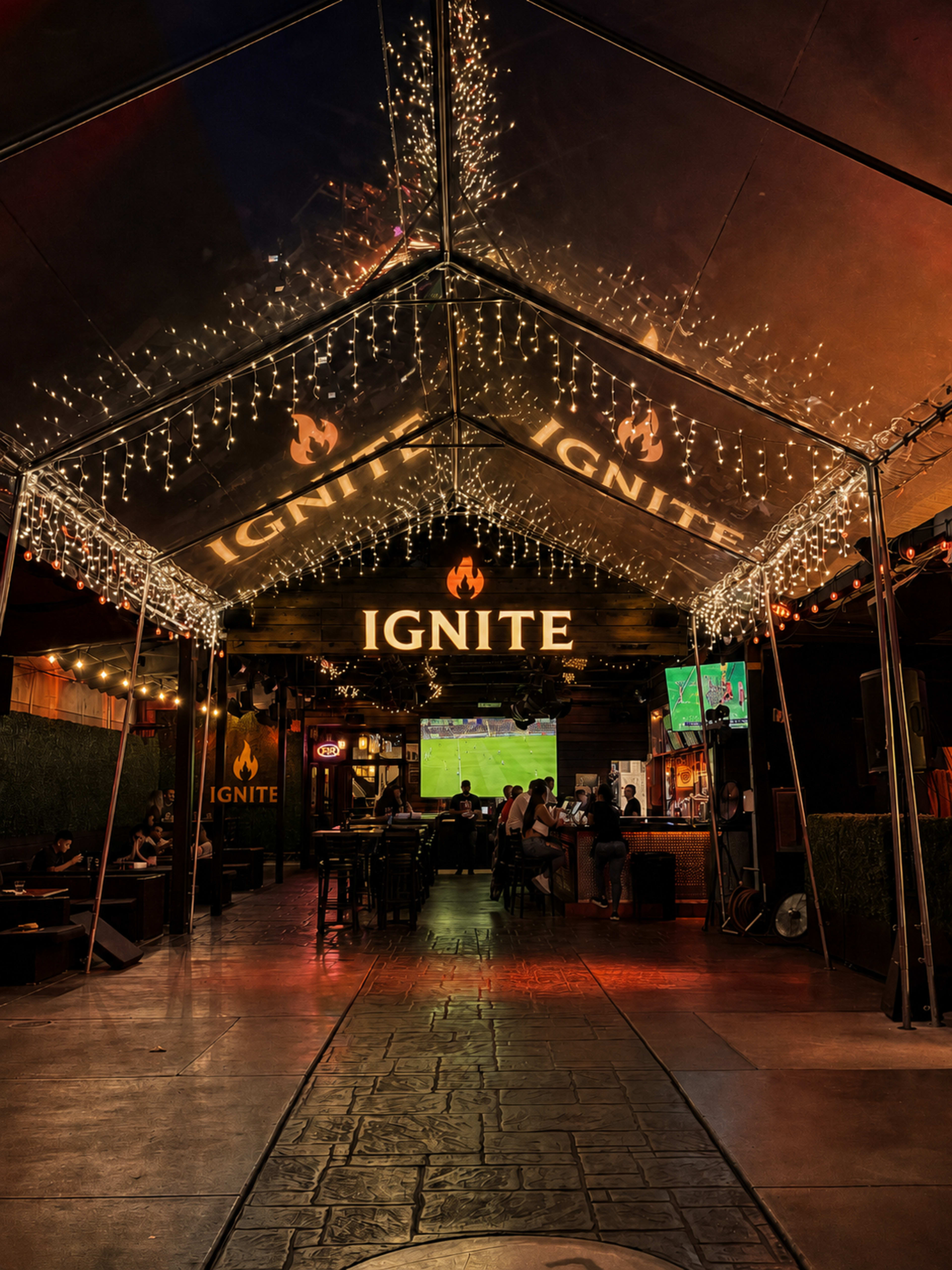 The image shows a lively bar area with a tented roof, decorated with string lights and featuring a large sign that reads "IGNITE."