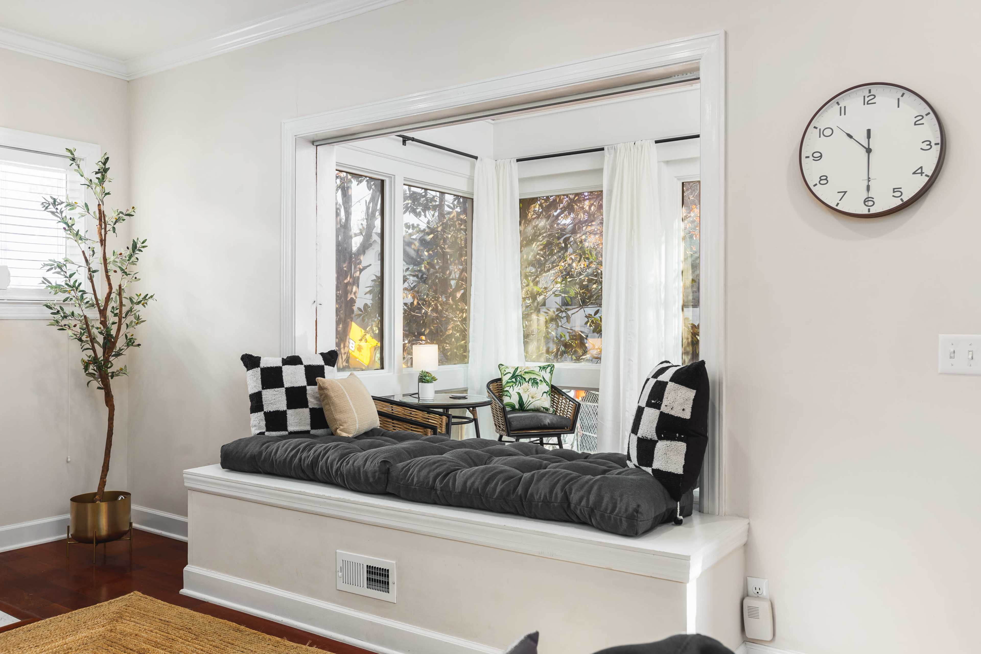 A cozy nook features a cushioned bench with checkered pillows, overlooking a sunlit room with large windows.
