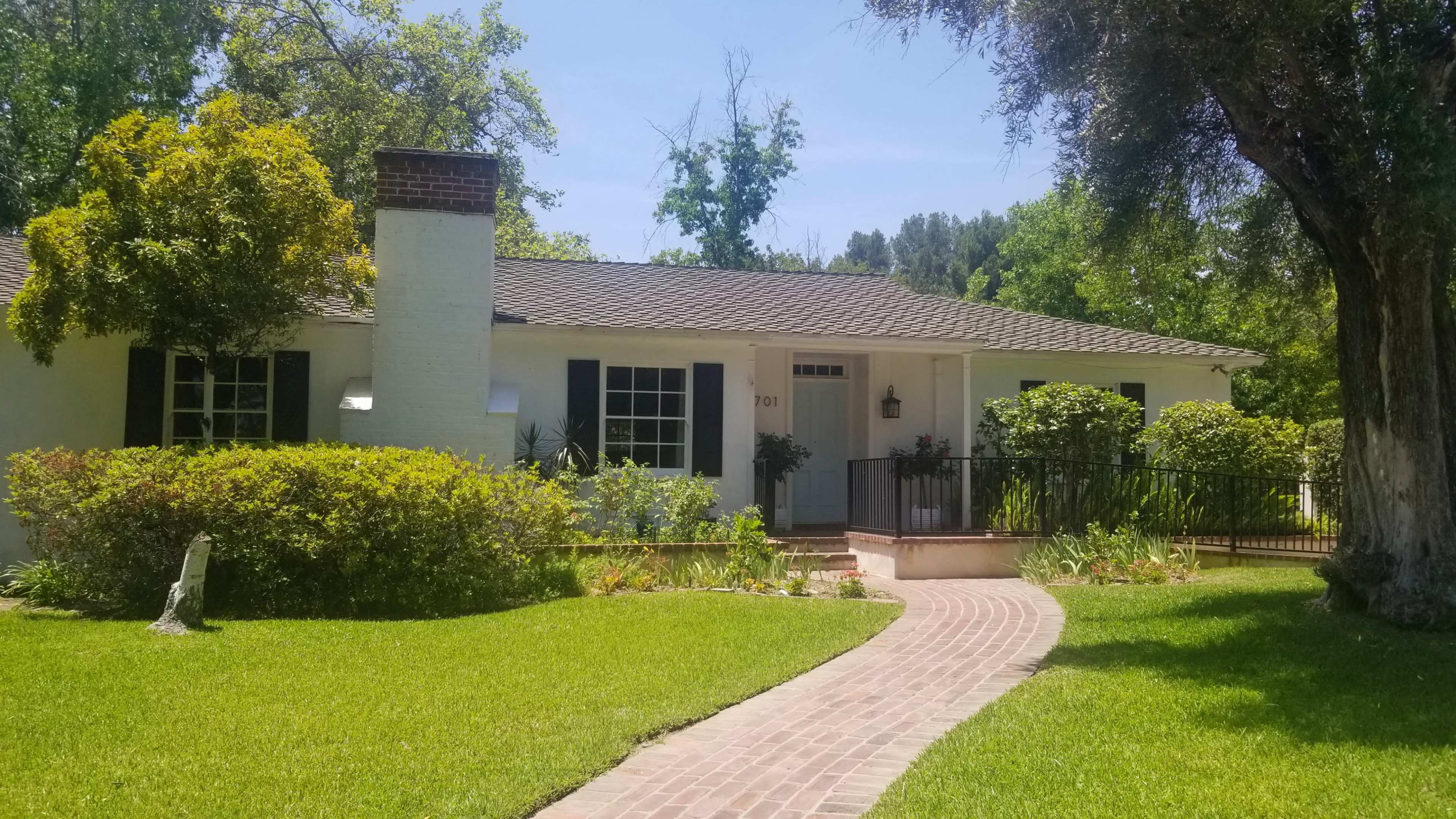A single-story, white house with black shutters and a brick walkway is surrounded by lush greenery and trees.