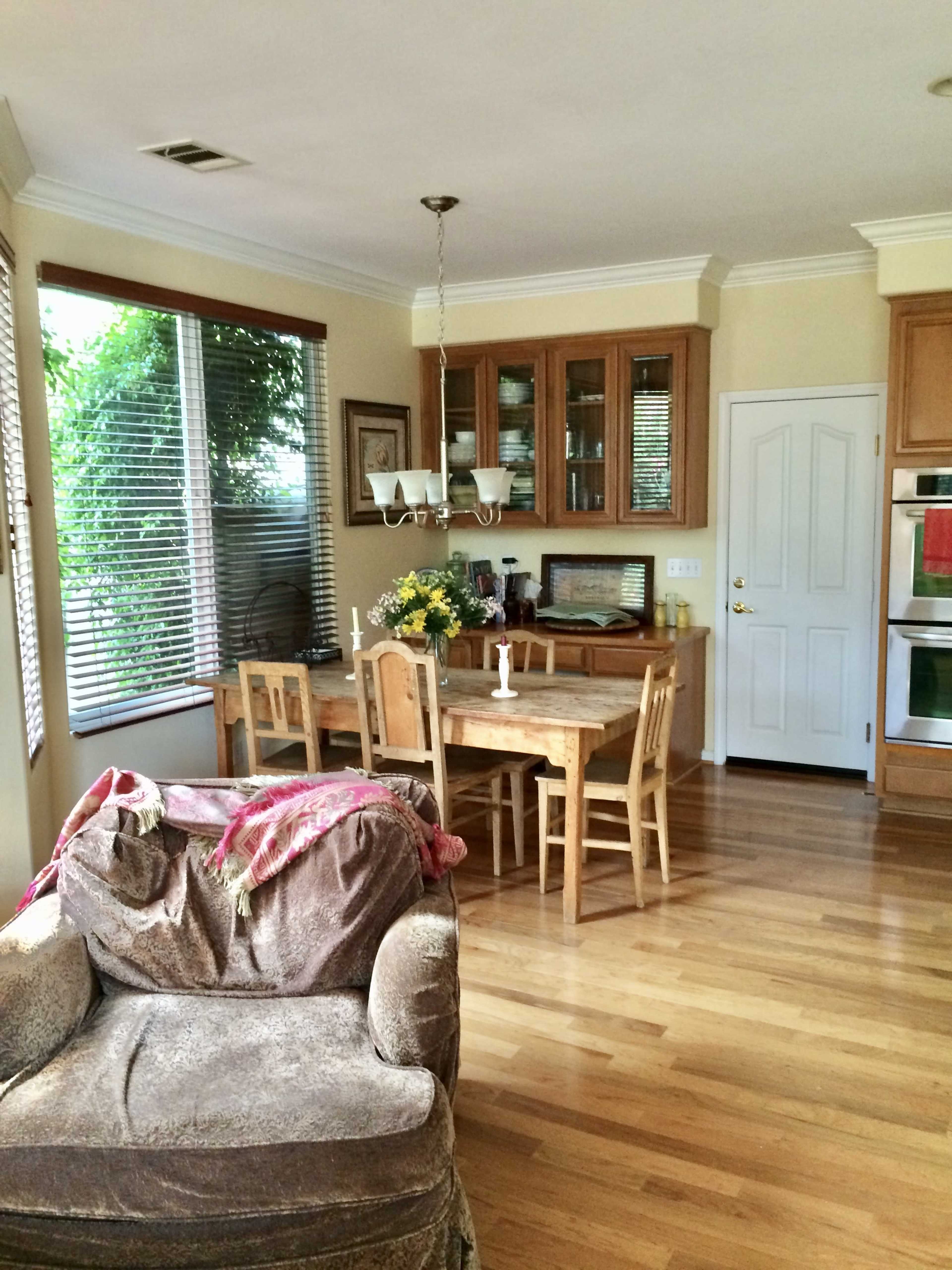A dining area with a wooden table and chairs, surrounded by large windows, and a kitchen visible in the background.