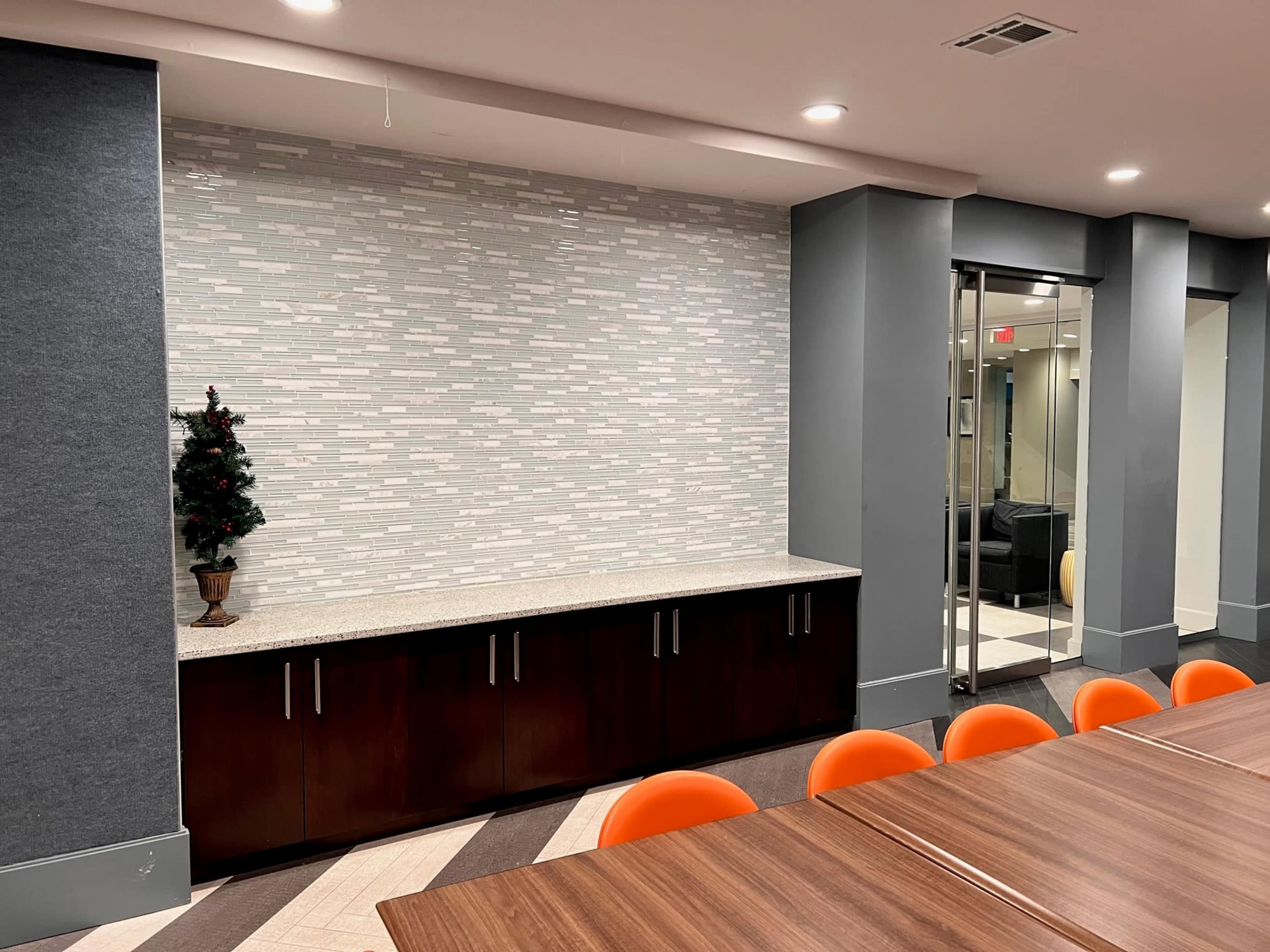 The image shows a modern meeting room with a long table, orange chairs, a dark wooden cabinet, and a textured gray wall with a decorative tree in the corner.
