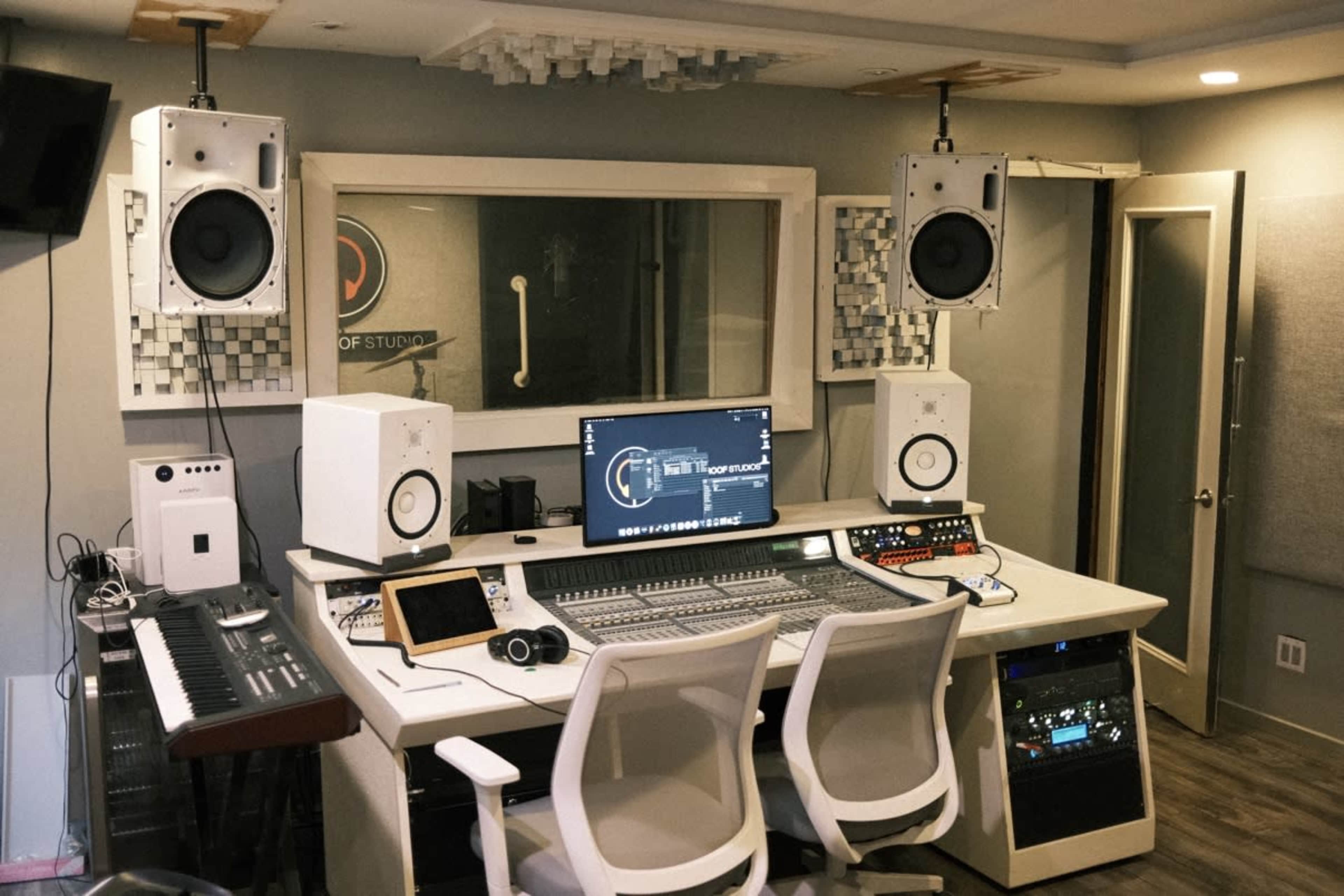 The image shows a recording studio with a central mixing desk, computer monitor, studio monitors on stands, and various music equipment arranged on a white desk.