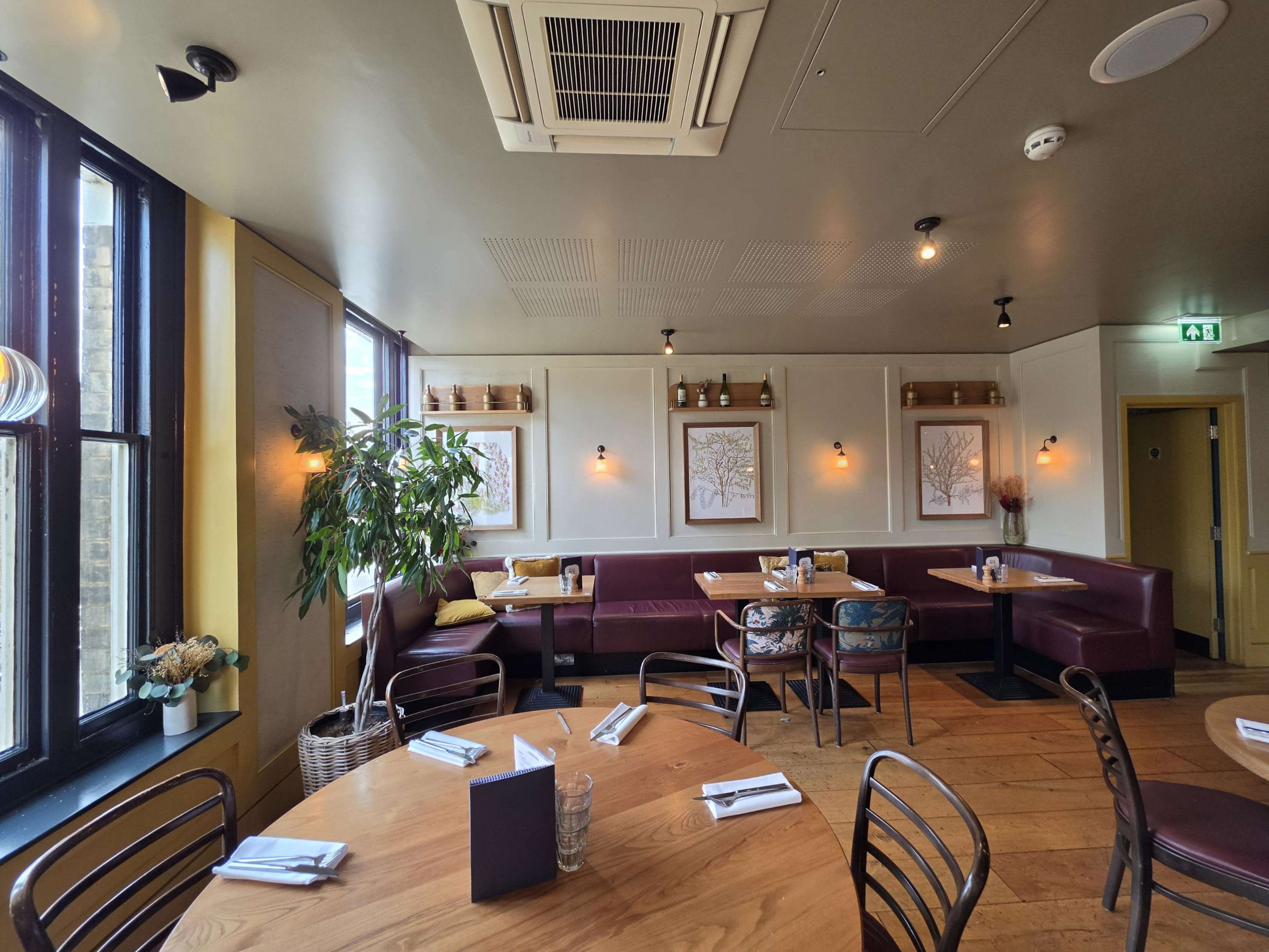 The image shows a cozy restaurant interior featuring wooden tables, burgundy upholstered seating, framed art on the walls, and a potted plant in the corner.
