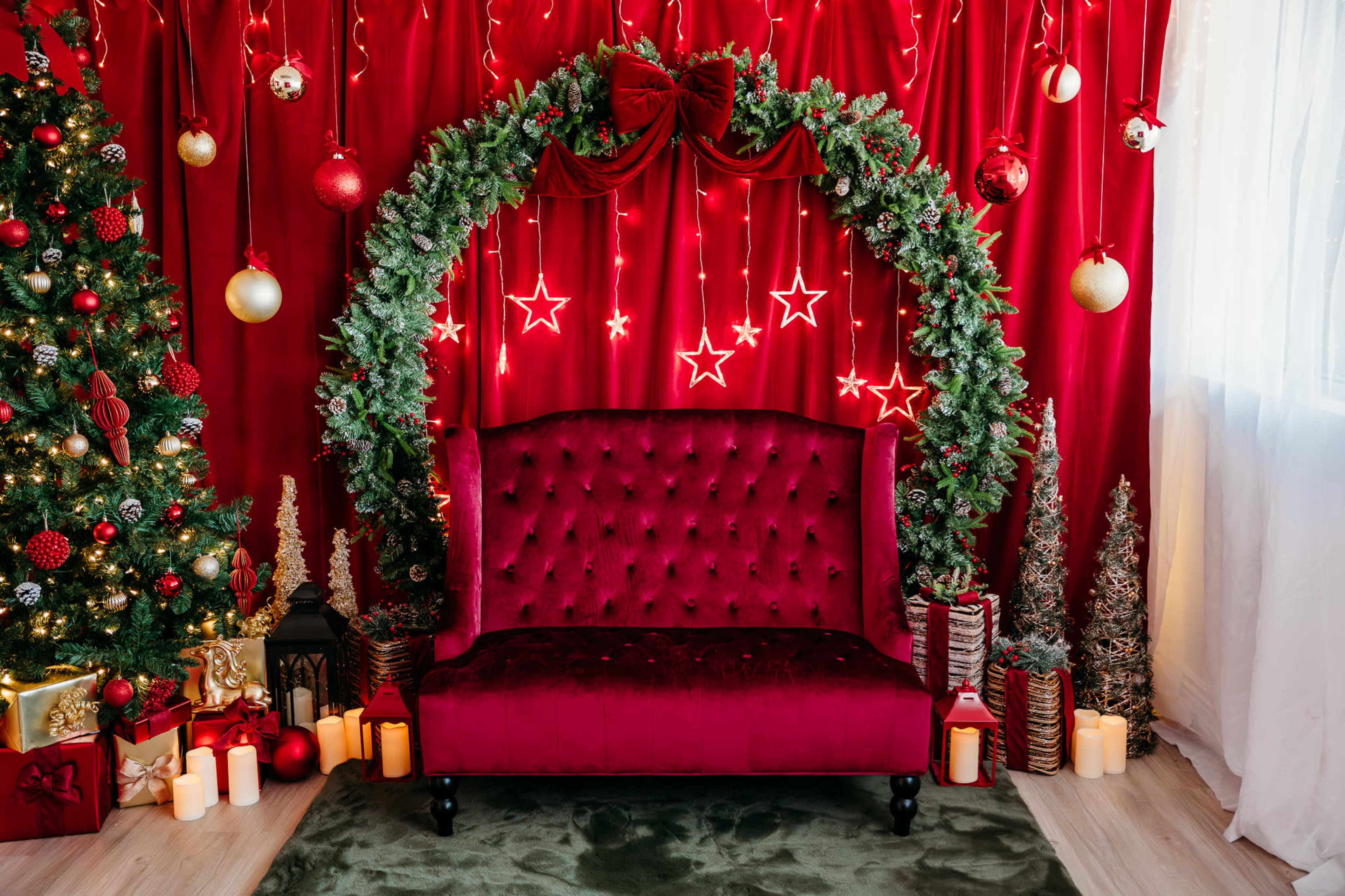 The scene features a festive red backdrop with a decorated Christmas tree, a wreath adorned with lights and ornaments, and a plush red couch surrounded by candles and holiday decorations.