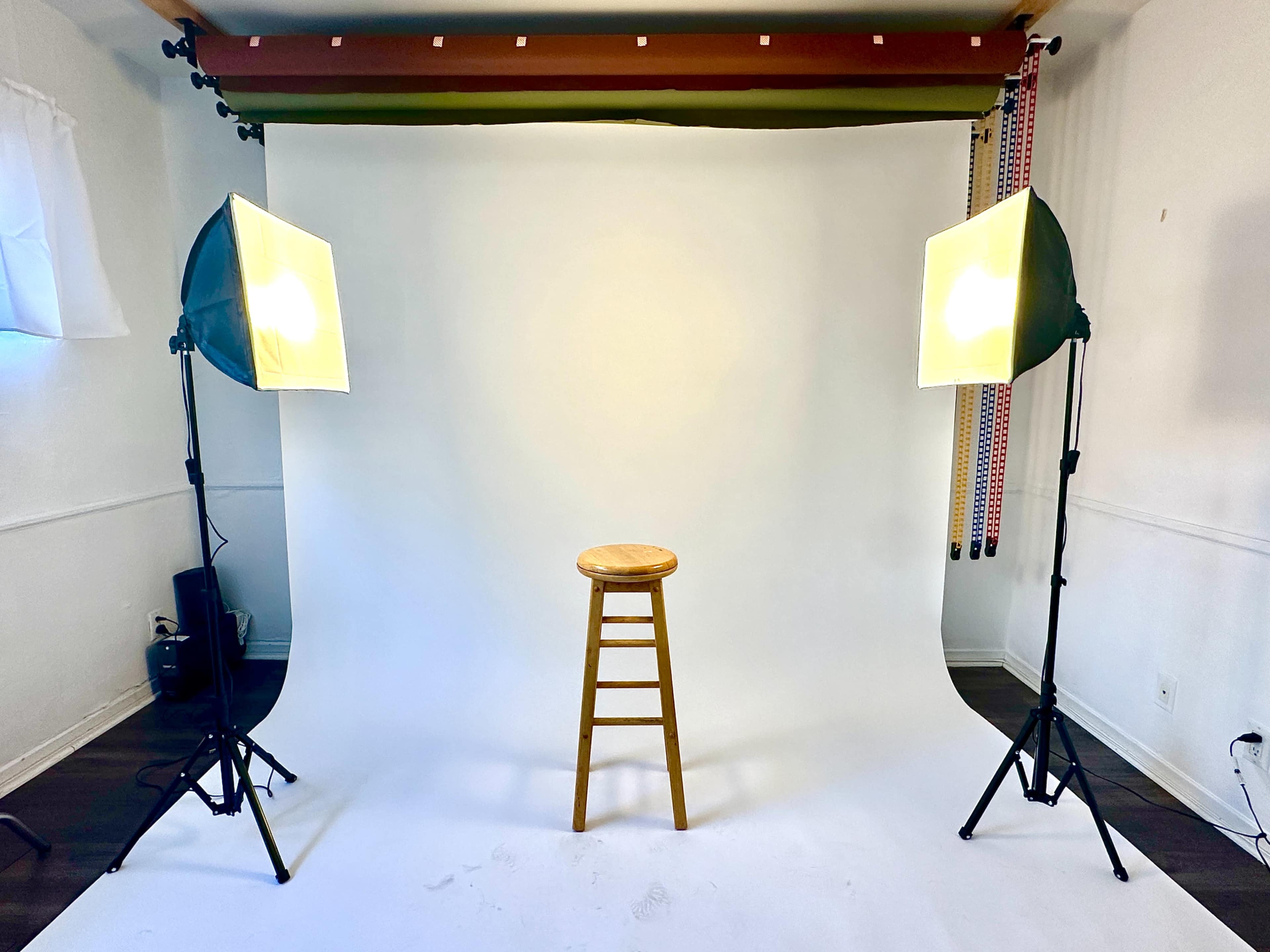 A wooden stool is positioned in front of a plain white backdrop, flanked by two studio lights on tripods.