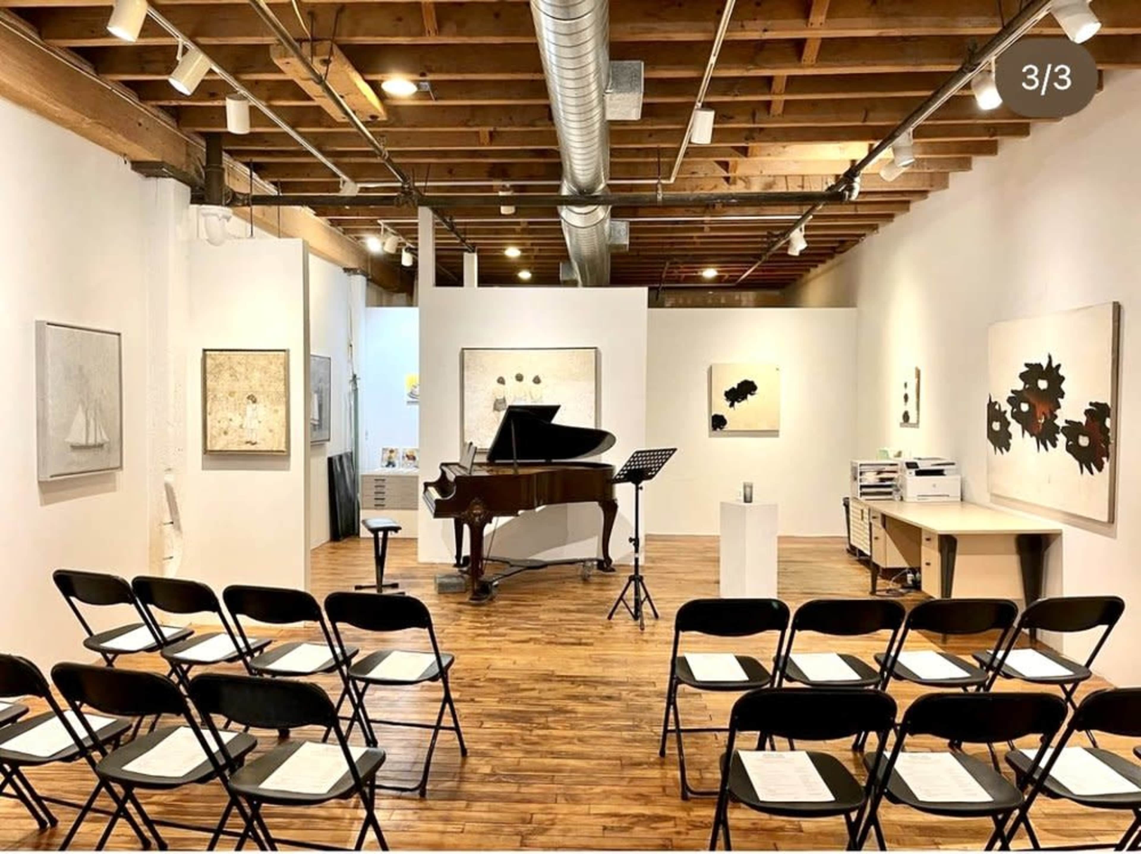 A music performance space with a grand piano in the center, surrounded by rows of black chairs and art displayed on the walls.
