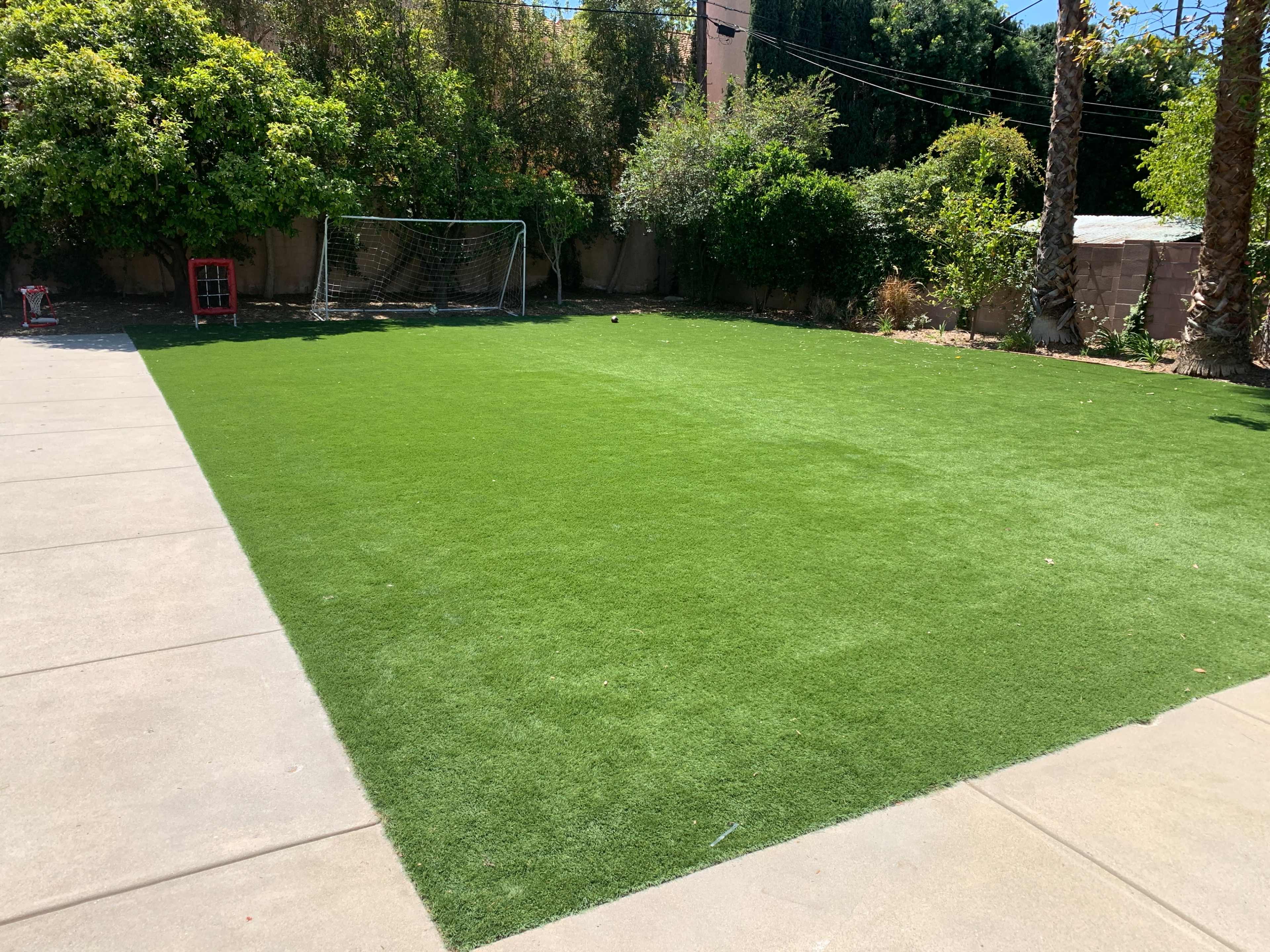 A manicured artificial turf lawn with a soccer goal in the background, surrounded by greenery and palm trees.