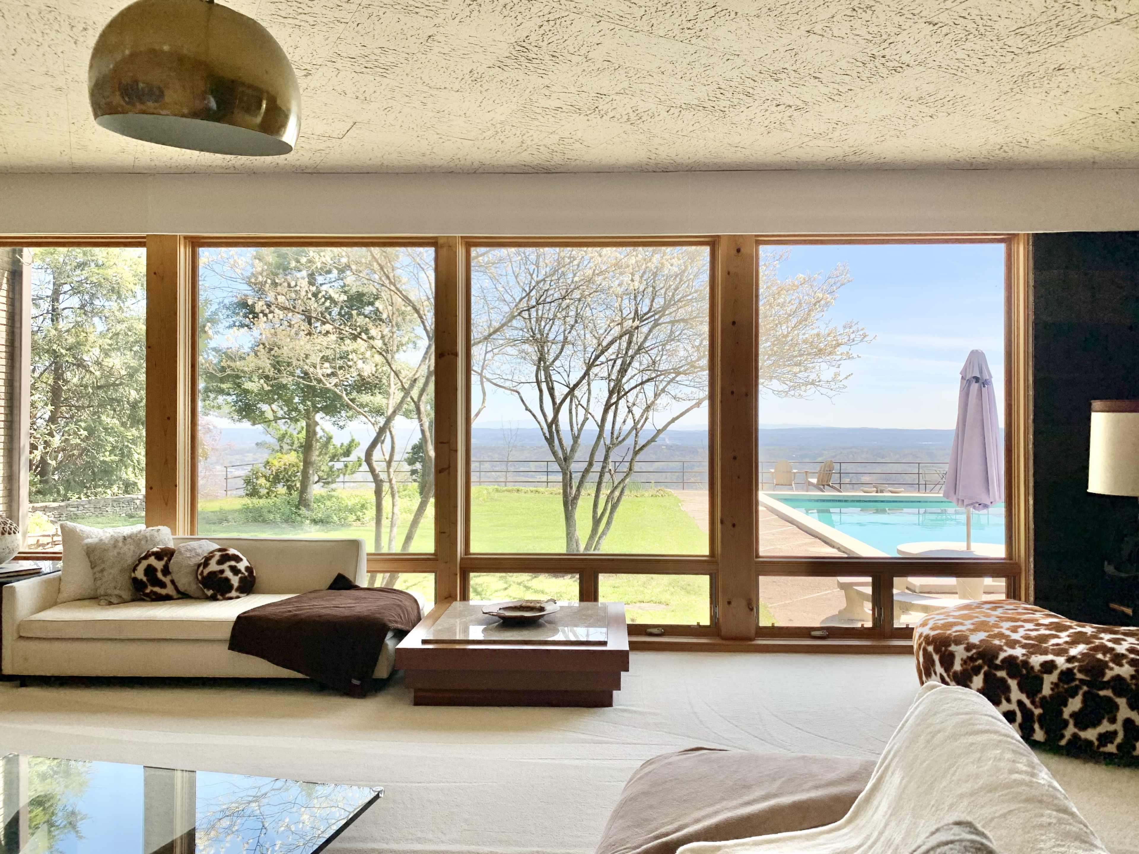 A spacious living room features large windows overlooking a pool and landscaped yard with trees.