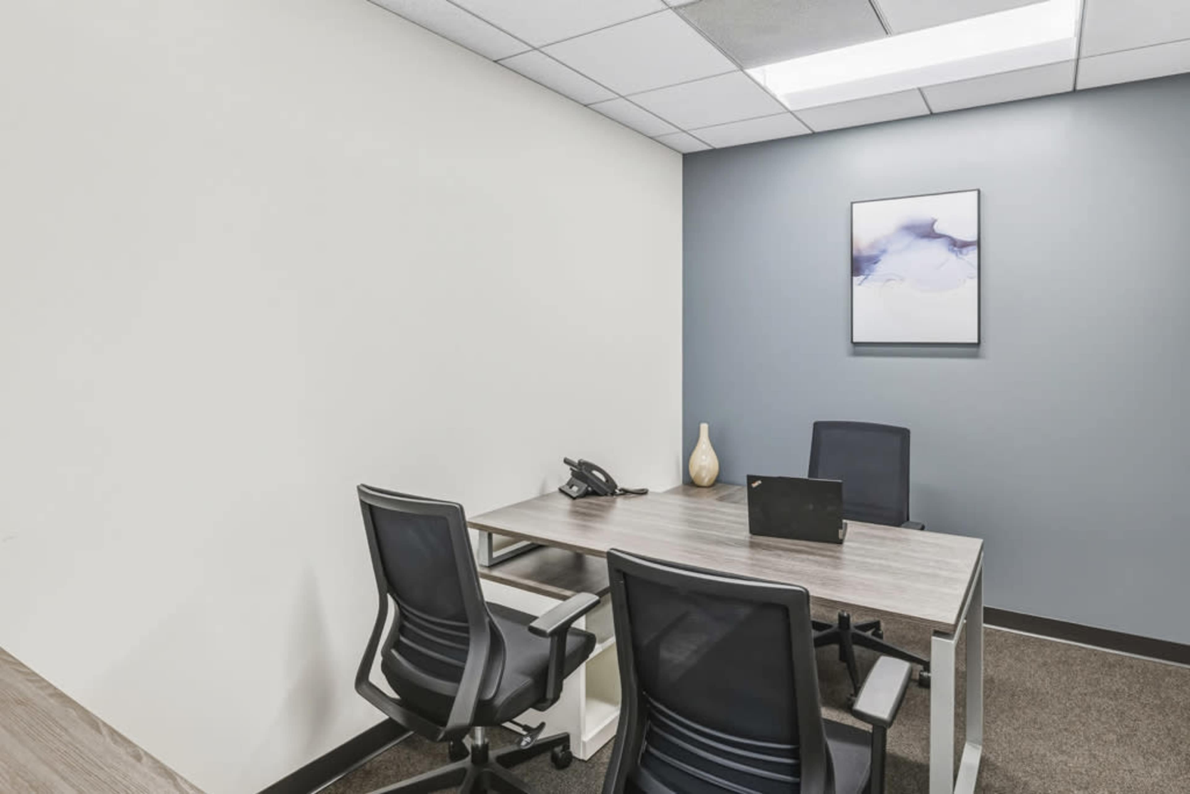The image shows a simple office space featuring a wooden desk with two office chairs, a phone, a laptop, and a decorative vase on the desk, with a painting on the wall.
