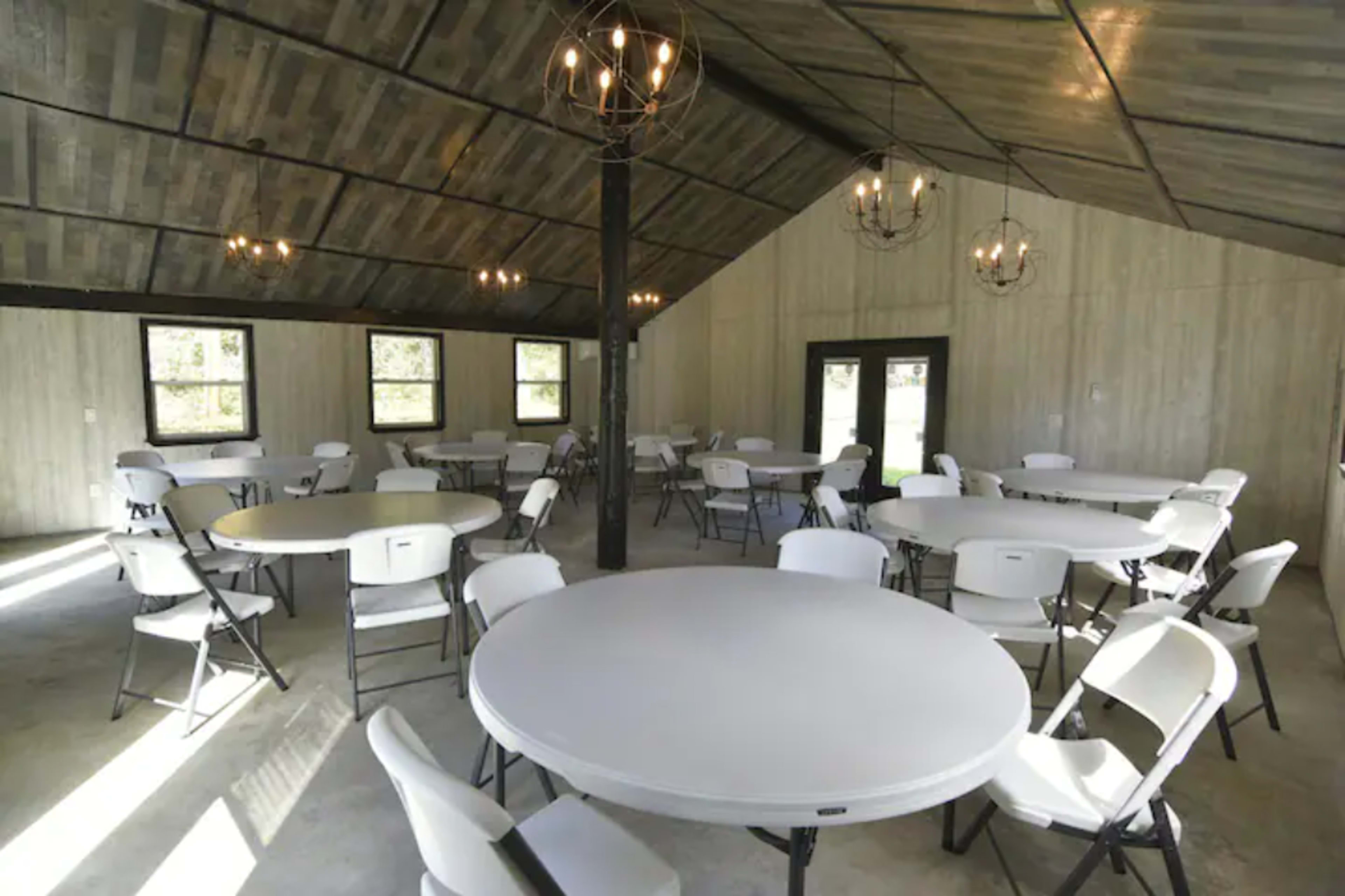 A spacious indoor venue features round tables surrounded by folding chairs under a high, vaulted ceiling with pendant lighting.