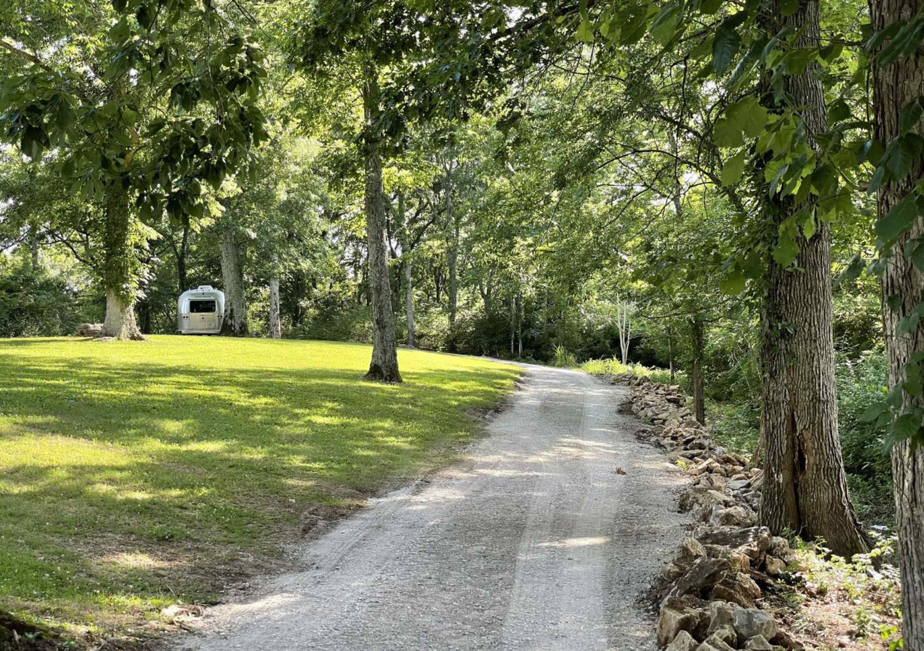 A gravel path winds through a green landscape lined with trees, leading to a small trailer in the distance.
