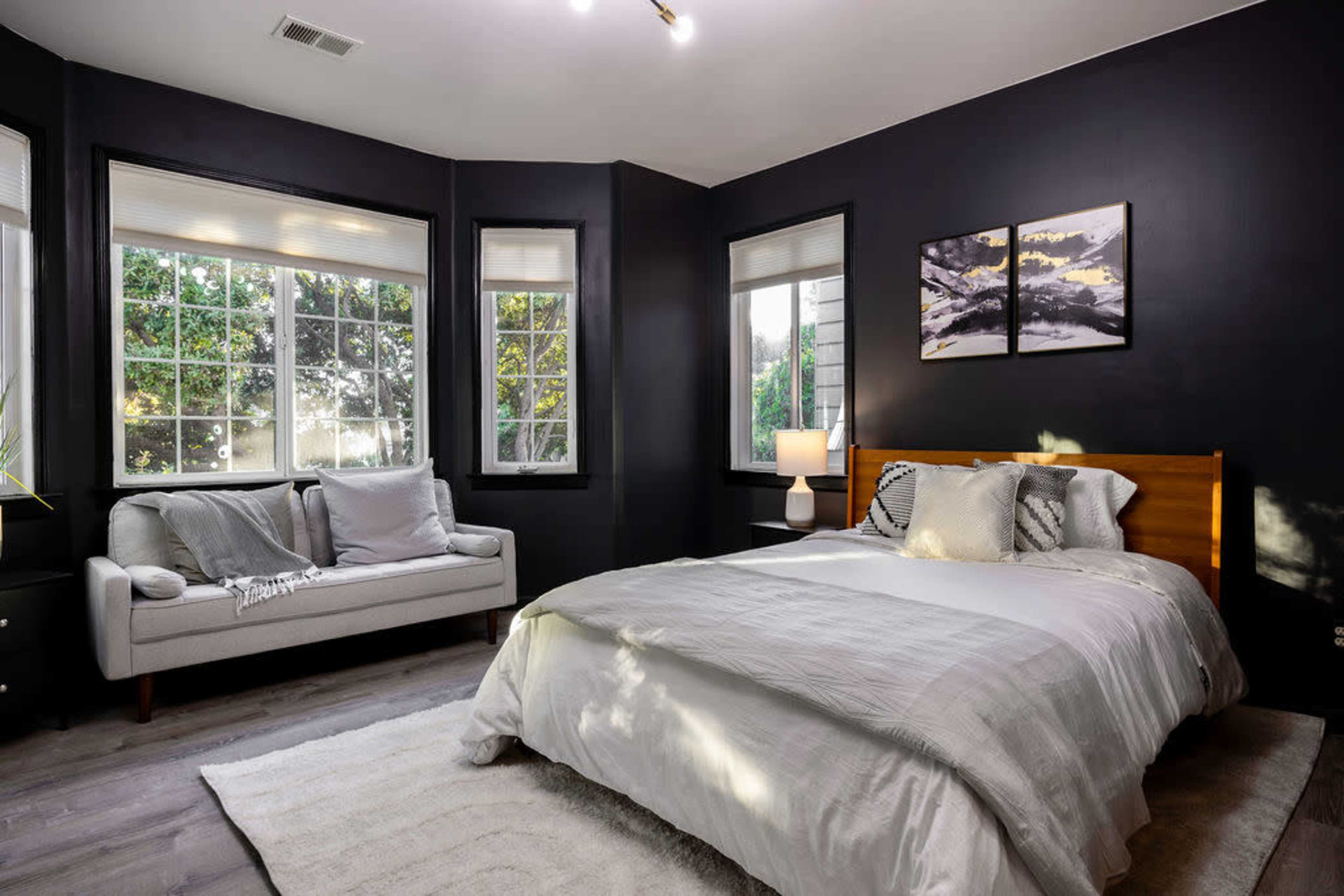A modern bedroom features a bed with light bedding, a white sofa, and large windows with natural light.