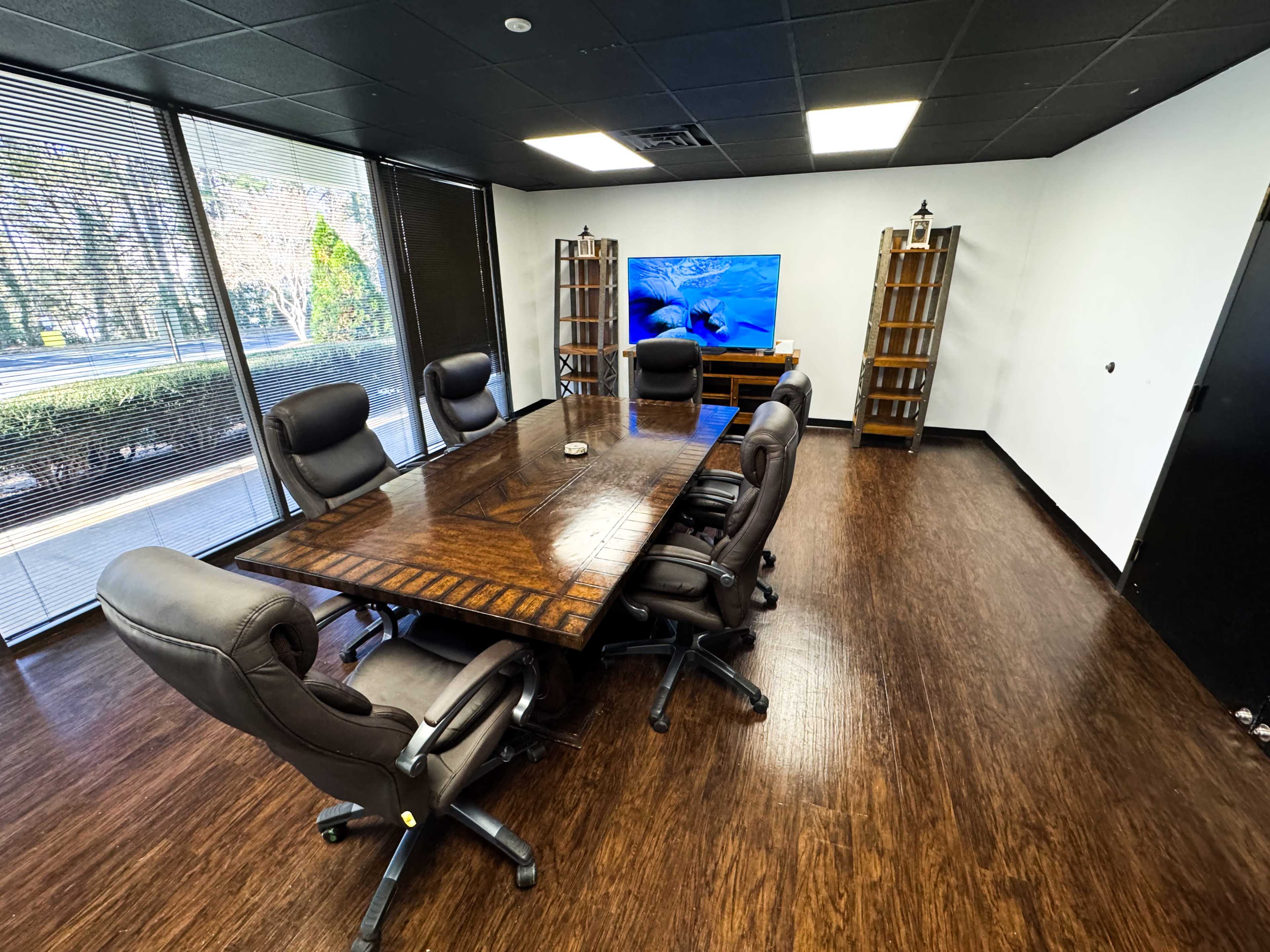 A conference room features a large wooden table surrounded by six leather chairs, with bookshelves and a television on the wall.