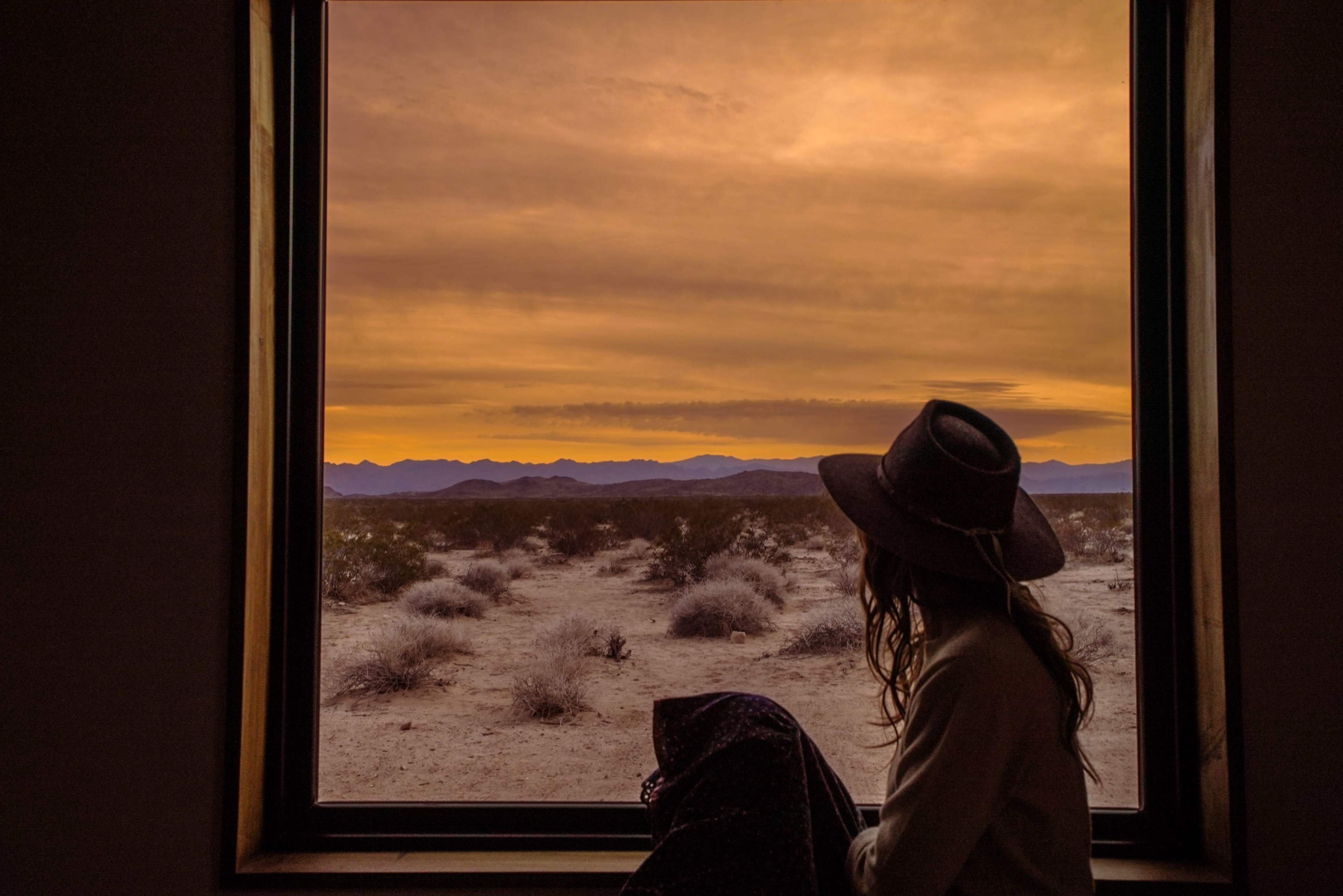 A person sits by a large window, gazing out at a desert landscape under an orange sky at sunset.