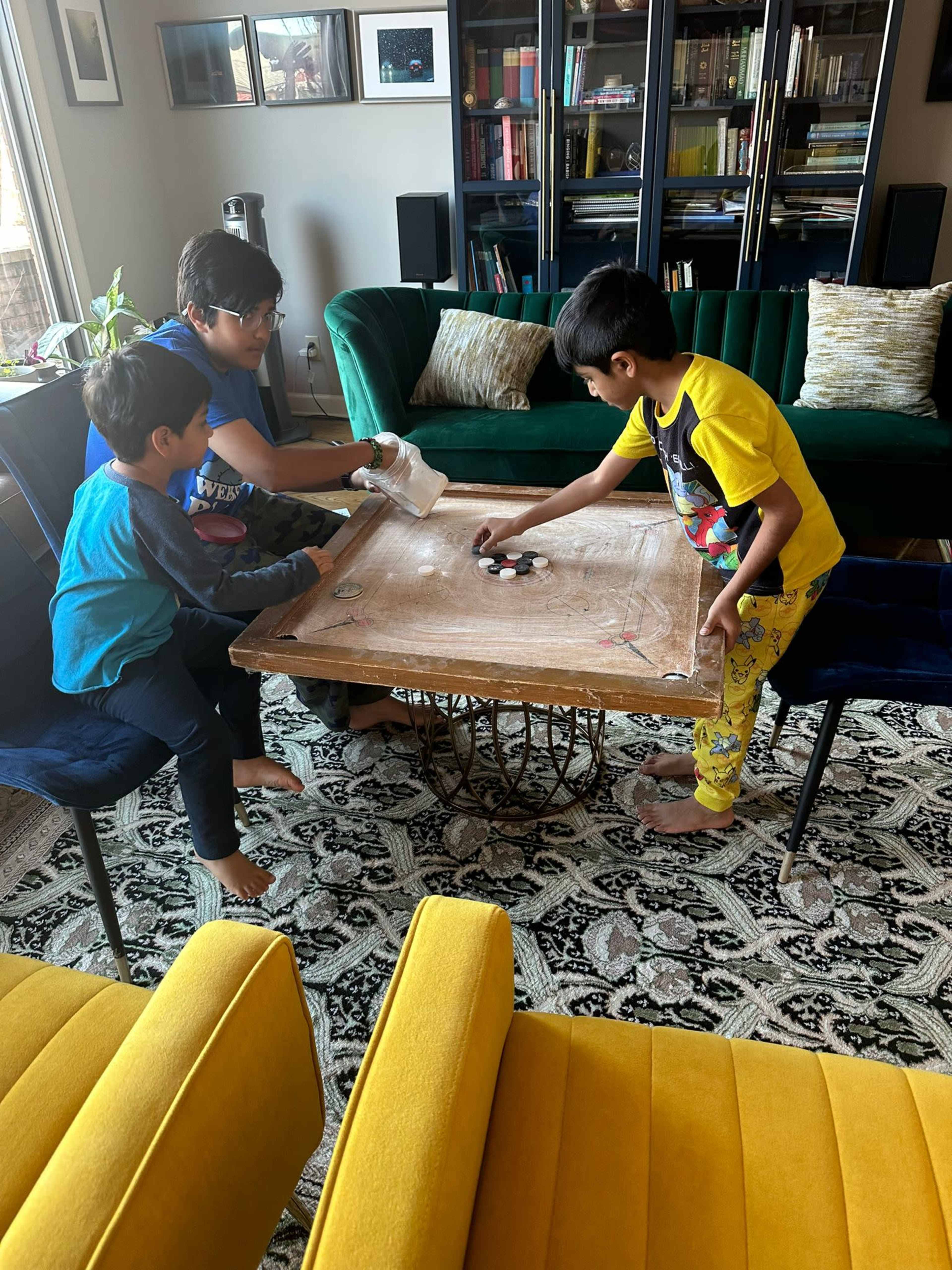 Three children are playing on a coffee table in a living room, with board game pieces scattered on the table and colorful chairs in the background.