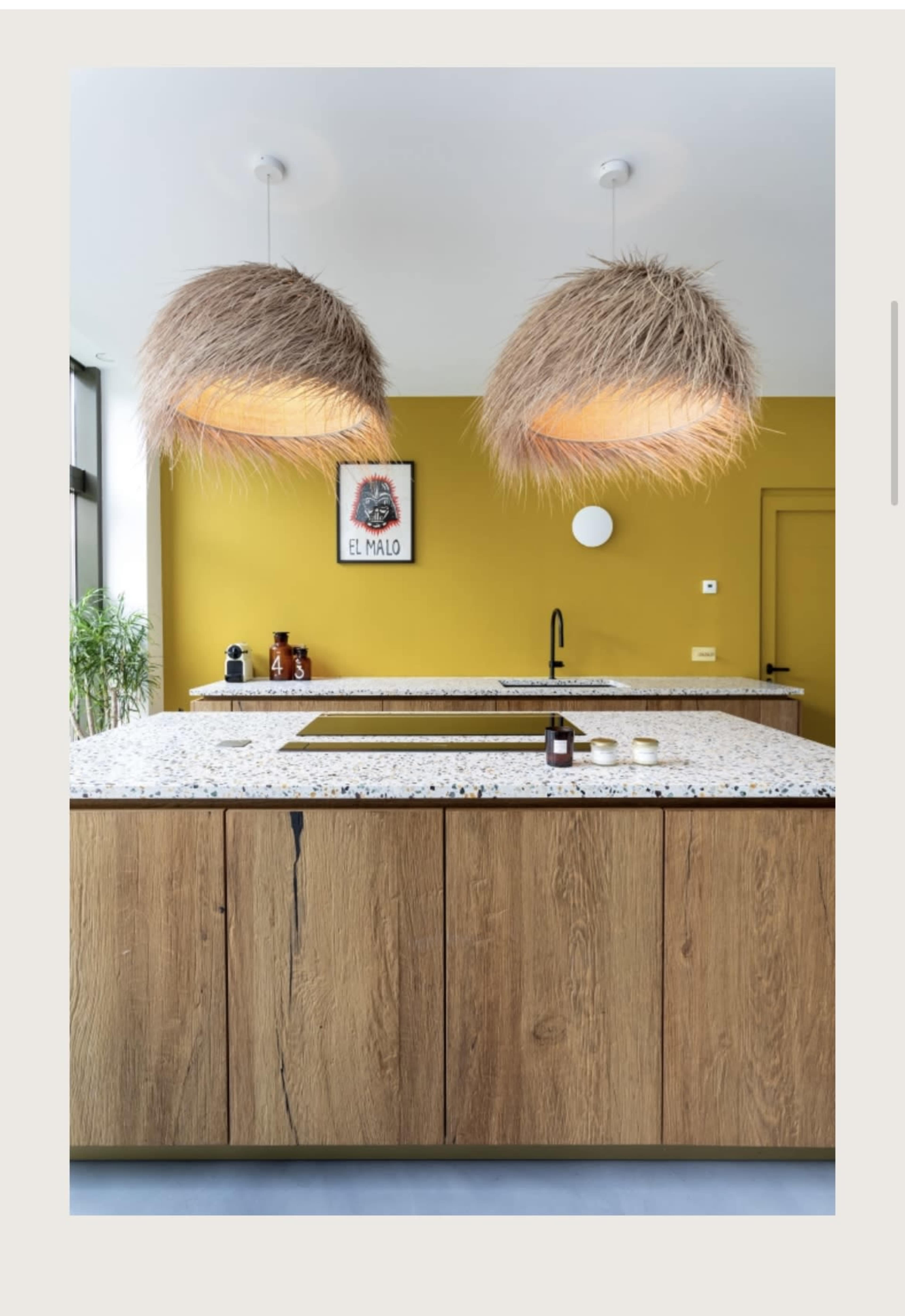 A modern kitchen with a yellow wall, two large pendant lights made of straw, and a central island with a speckled countertop.