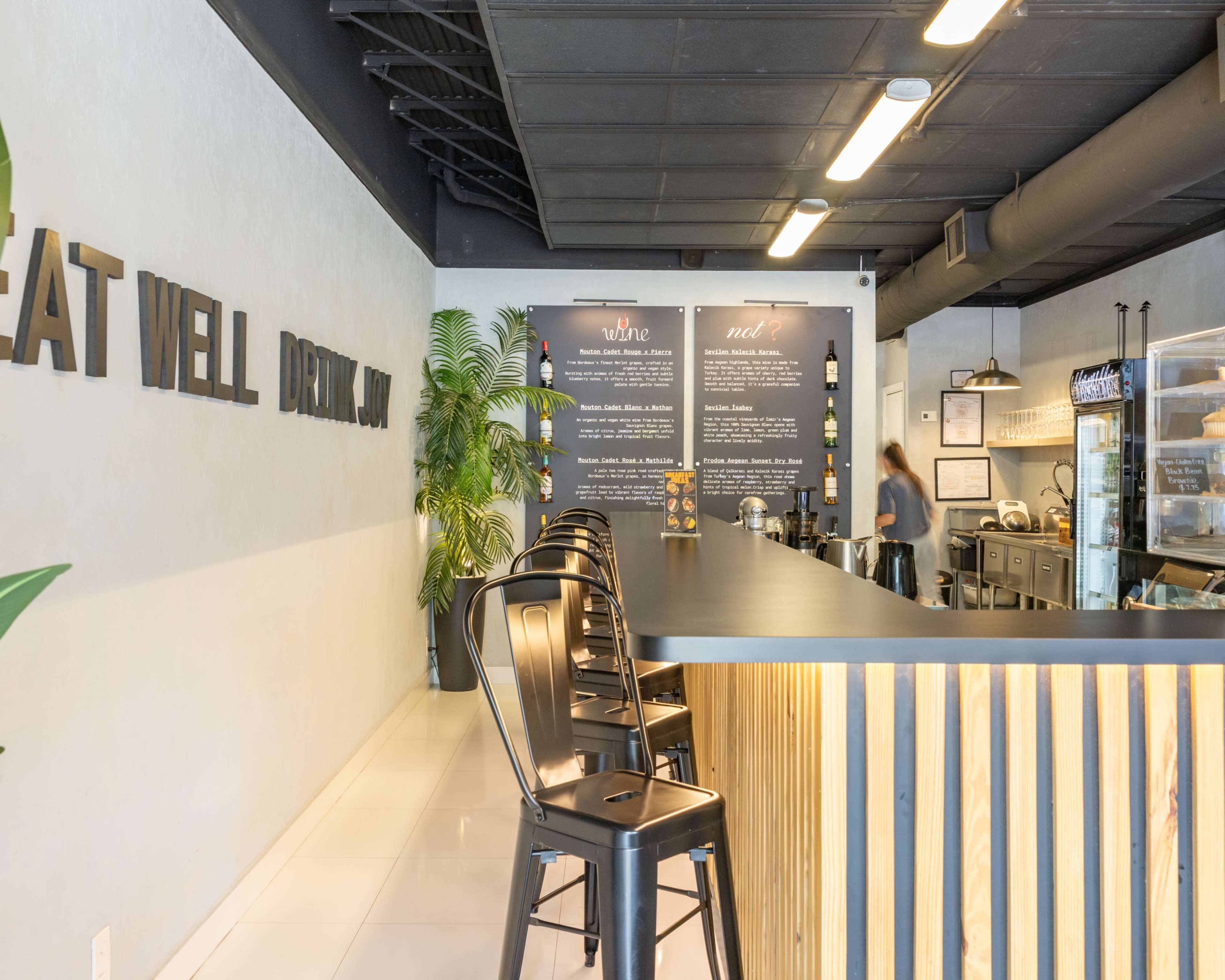 The image shows a clean, modern café interior with a long black counter, metal stools, and a wall featuring menu boards.