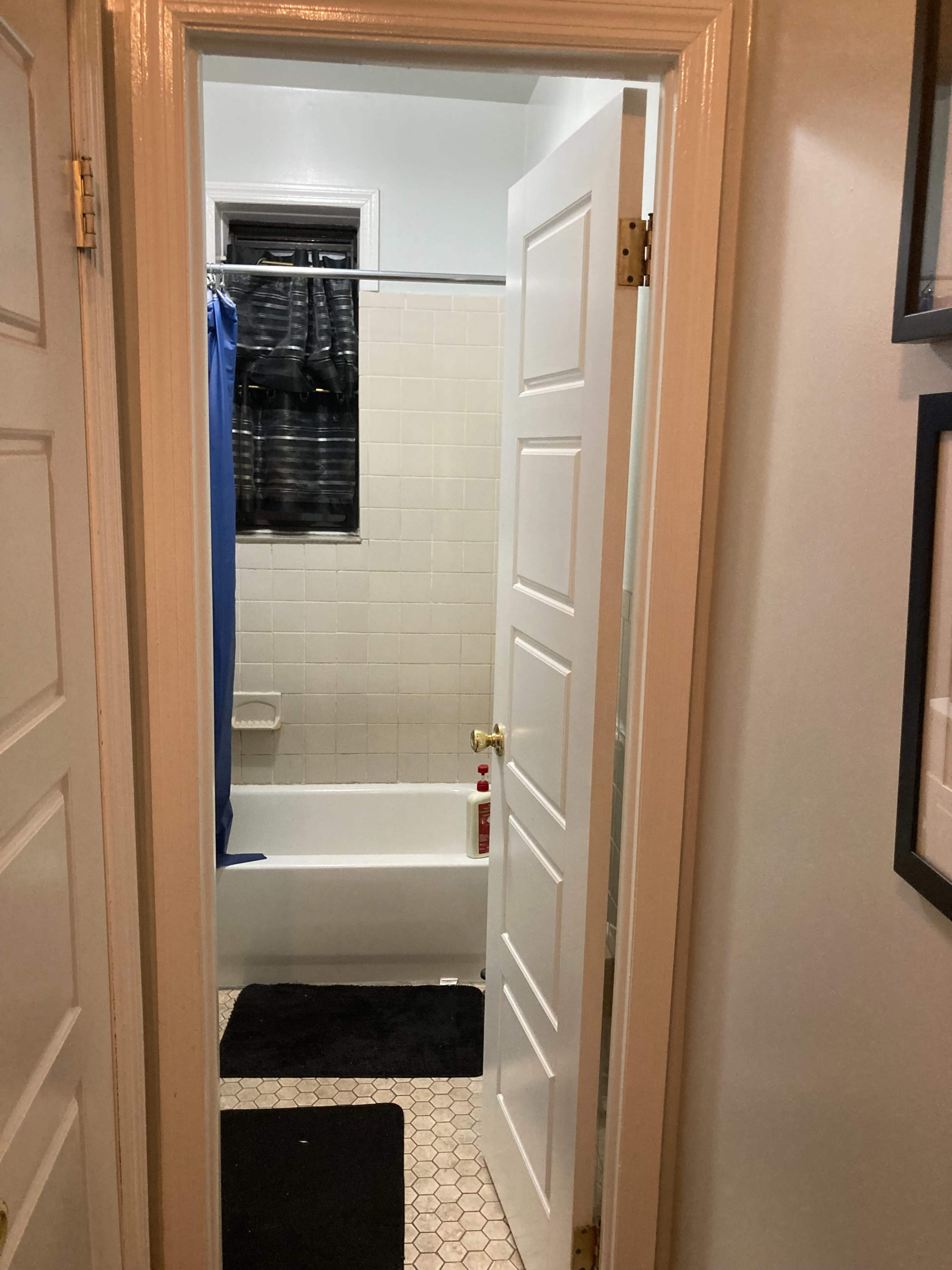 The image shows a bathroom doorway leading to a tub, with a blue shower curtain and black mats on the tiled floor.