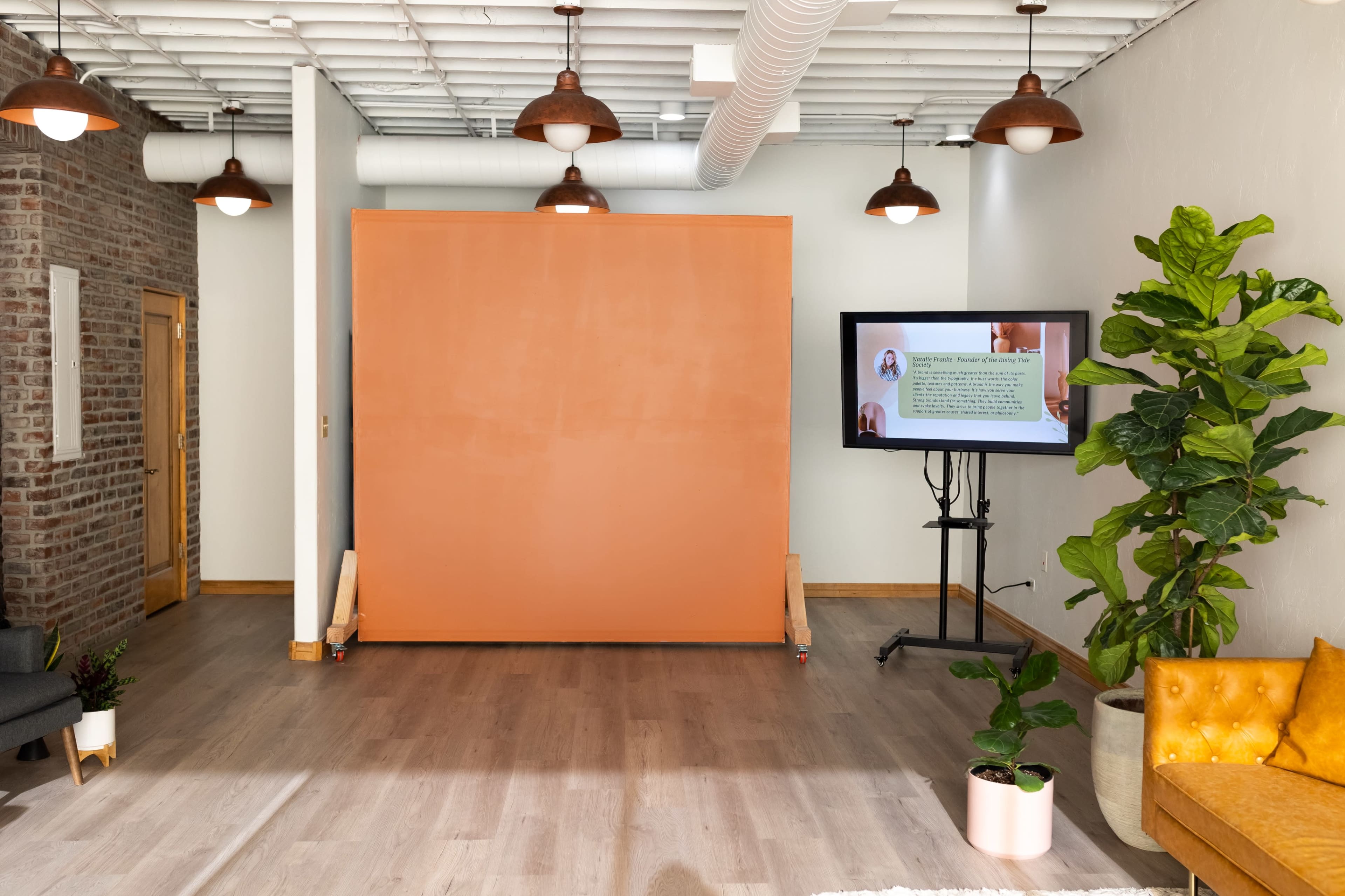 A bright interior space featuring an orange backdrop on wheels, a large television screen displaying text, and a potted plant, with seating arrangements in the corners.