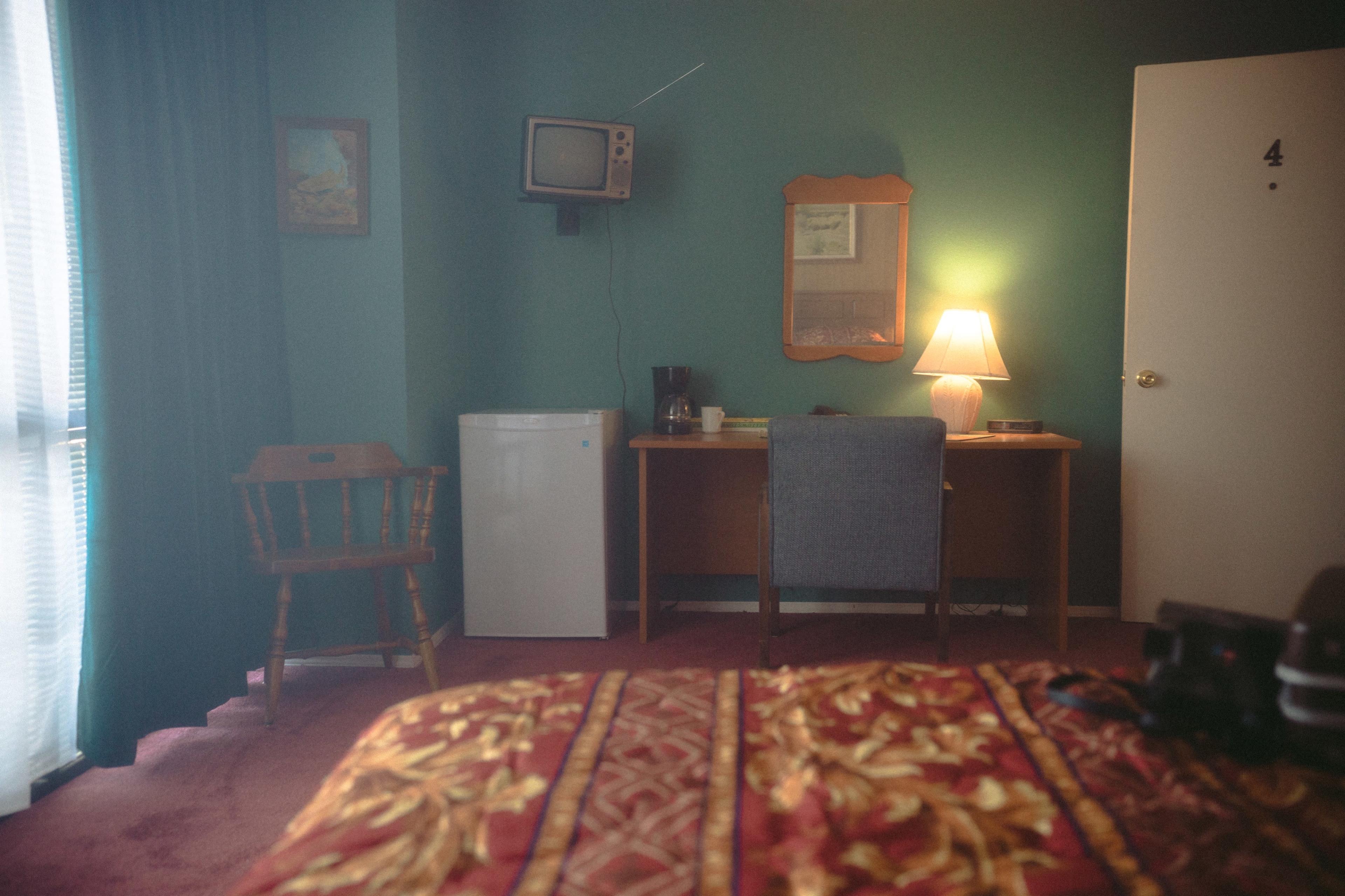 The image shows a small hotel room featuring a bed with patterned linens, a wooden desk with a chair, a mini fridge, an old television on the wall, a lamp, and a door with the number four.