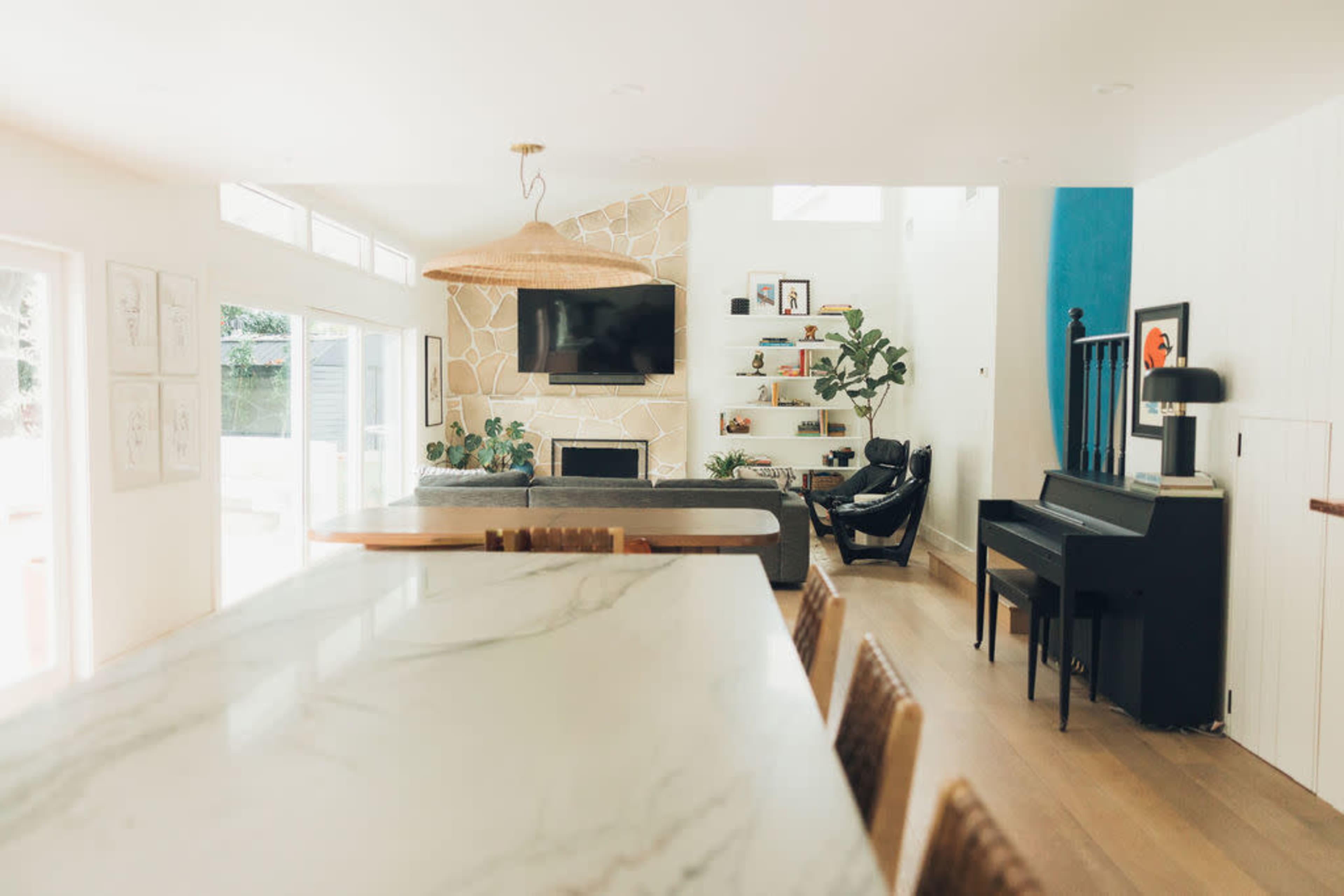 The image shows a bright, modern living space featuring a piano, a large television set, and a decorative stone wall, with natural light streaming in through large windows.
